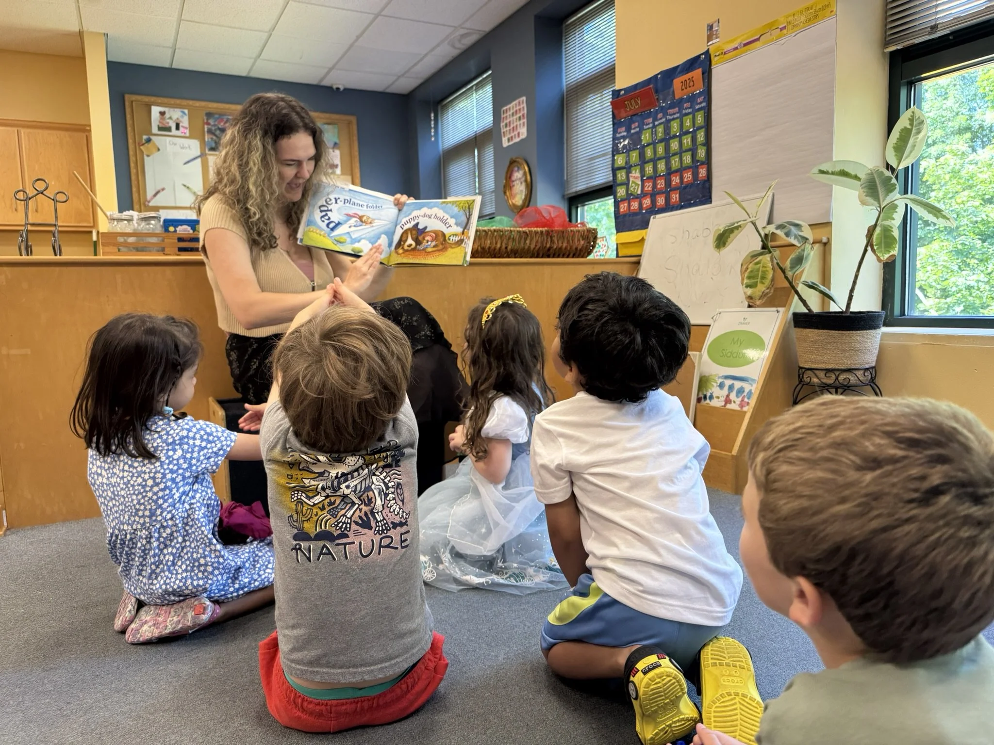 Children sitting on the floor in a classroom listening to a woman reading a picture book.