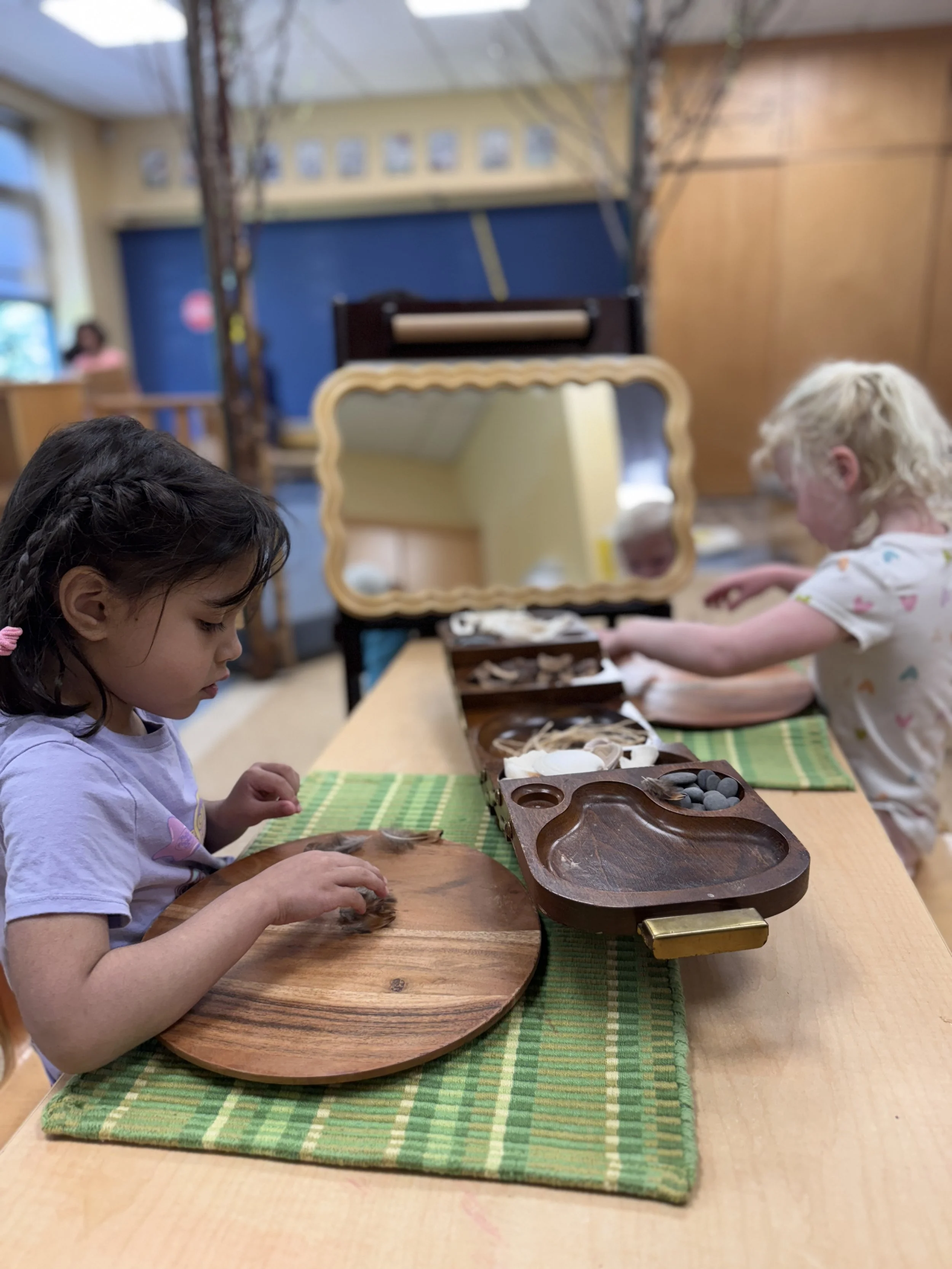 Two young girls at a wooden table with a green placemat, engaging in finger painting or arts and crafts, with various small items and wooden trays in front of them, and a mirror reflecting part of the room.