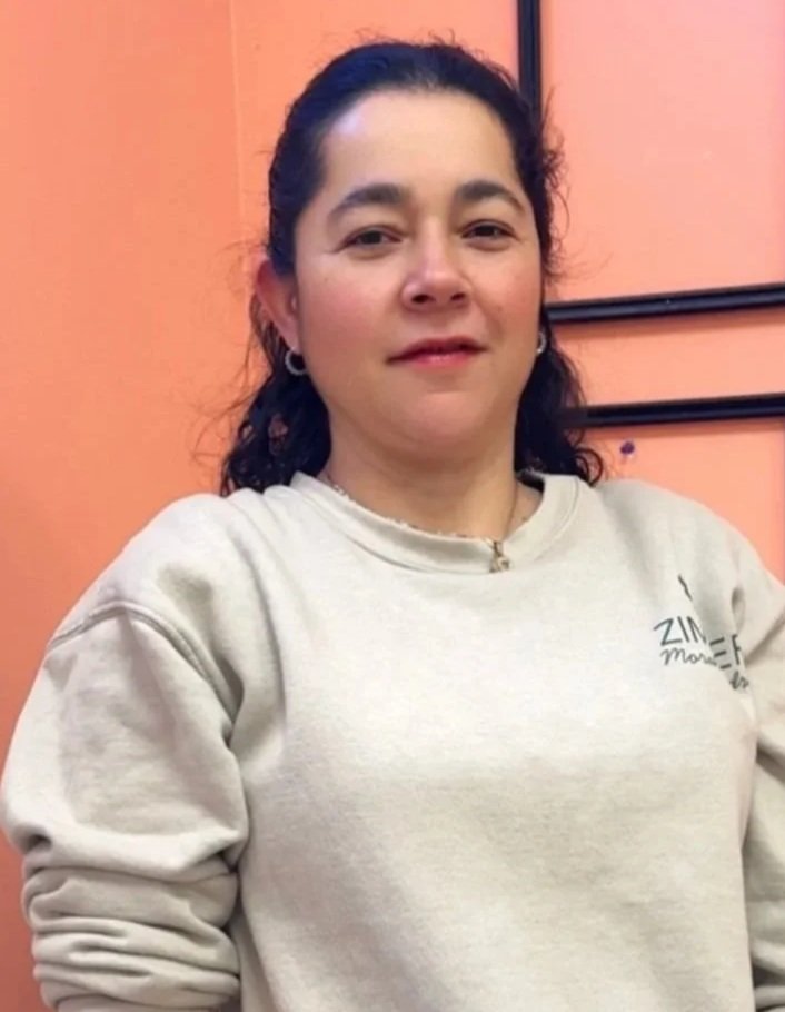 A woman with dark hair wearing a beige sweatshirt and small hoop earrings, standing in front of an orange wall.