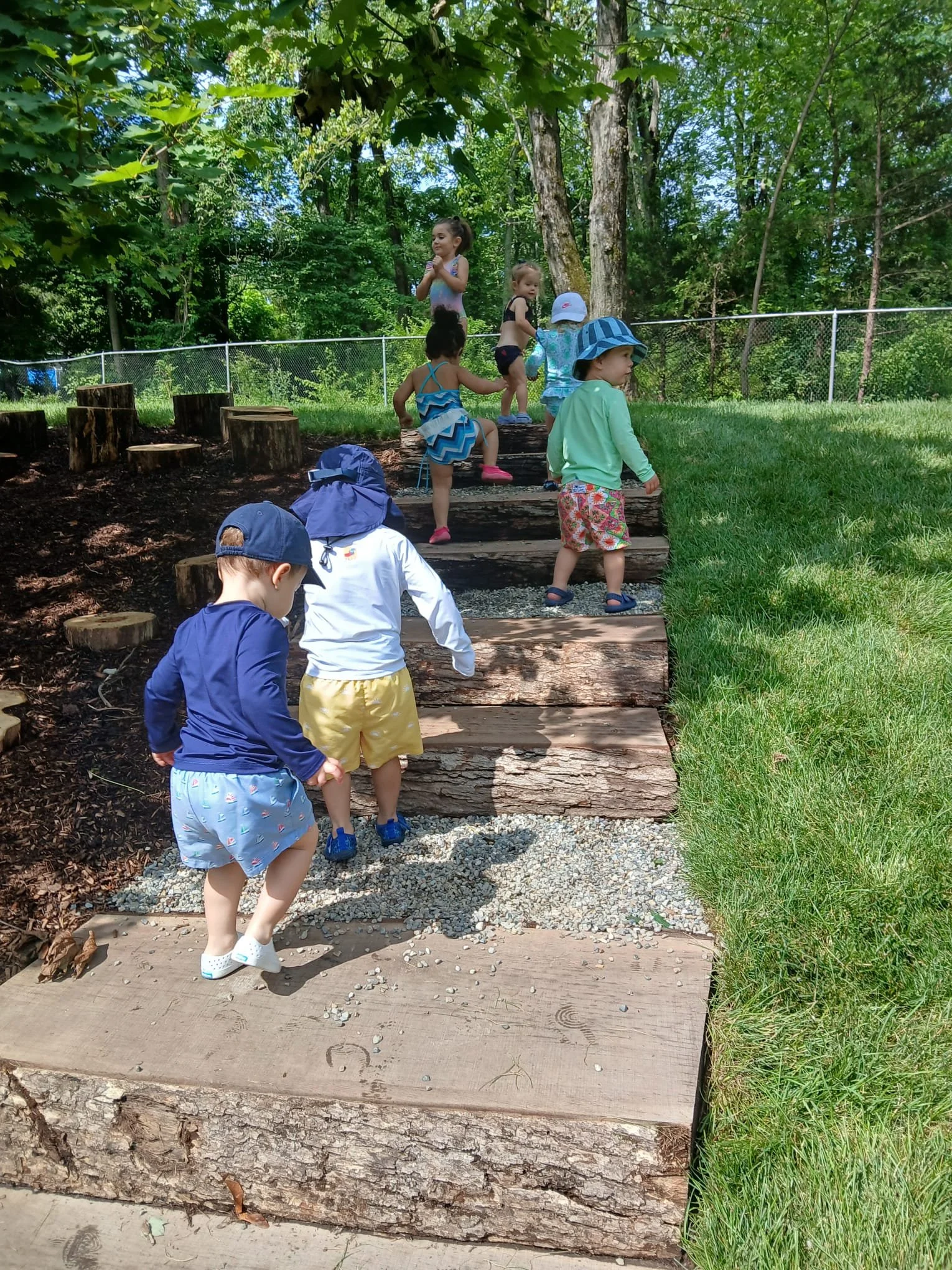 Children wearing hats and casual clothes climbing wooden steps outdoors in a wooded area with green trees and grass.