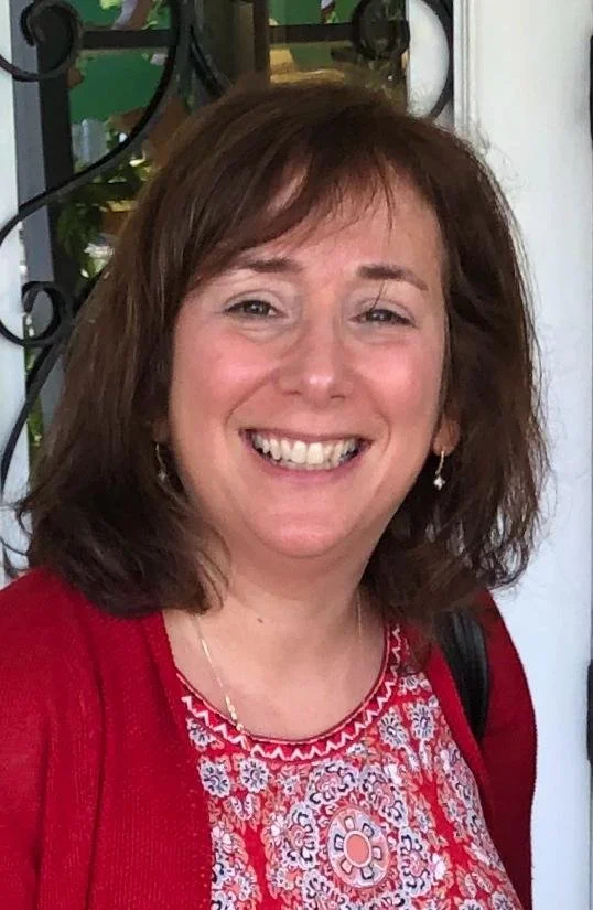 A smiling woman with shoulder-length brown hair wearing a red and white patterned blouse and earrings, standing in front of a white door with a black metal railing and green plants visible in the background.