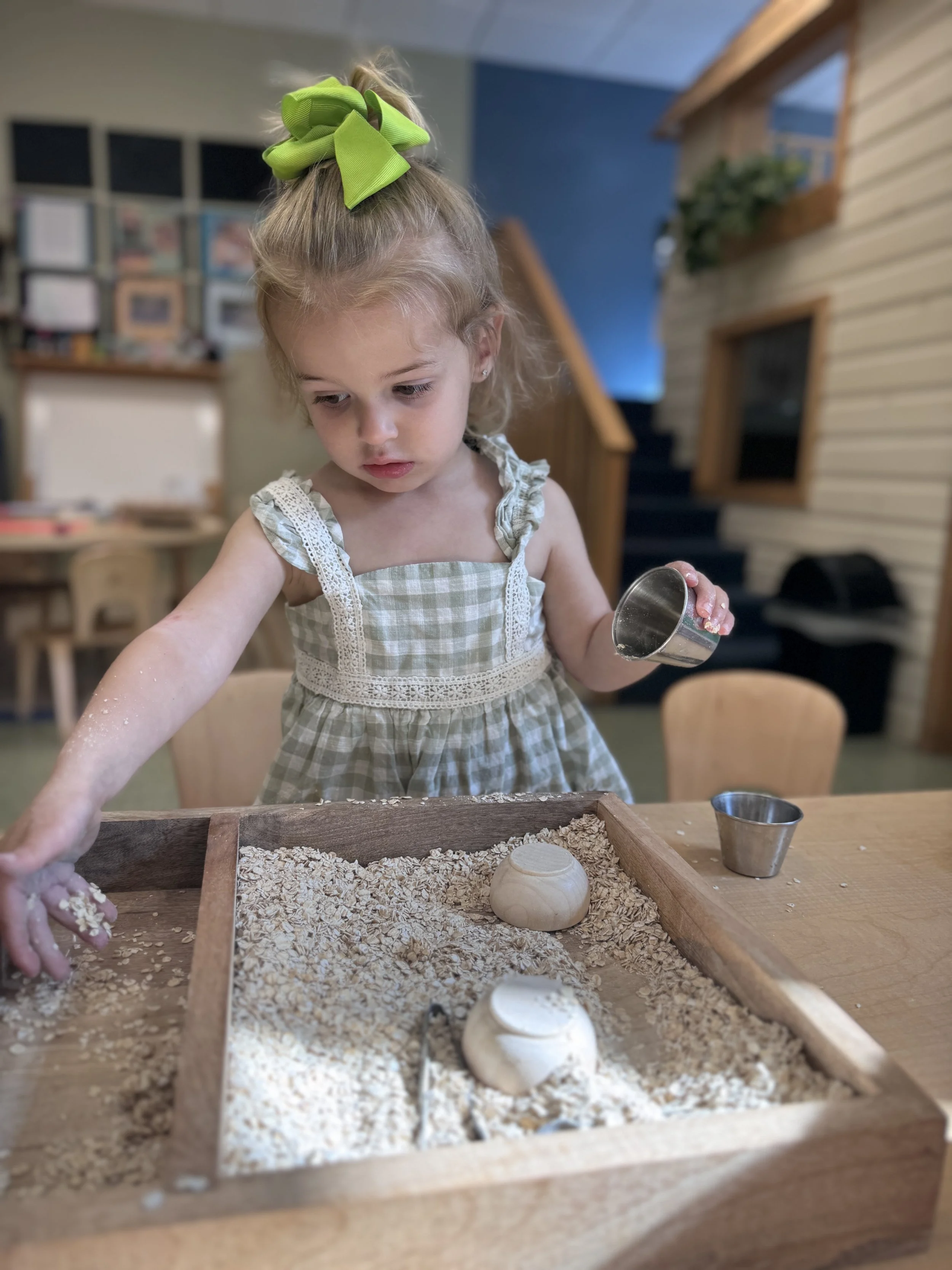 A young girl with a green bow in her hair playing with a sandbox filled with sand and small clay objects at a wooden table.