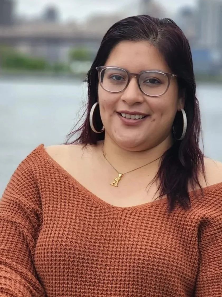 A woman with dark brown hair, glasses, and large hoop earrings, smiling outdoors by a body of water, wearing a brown knit sweater and gold necklace with a letter 'A' pendant.