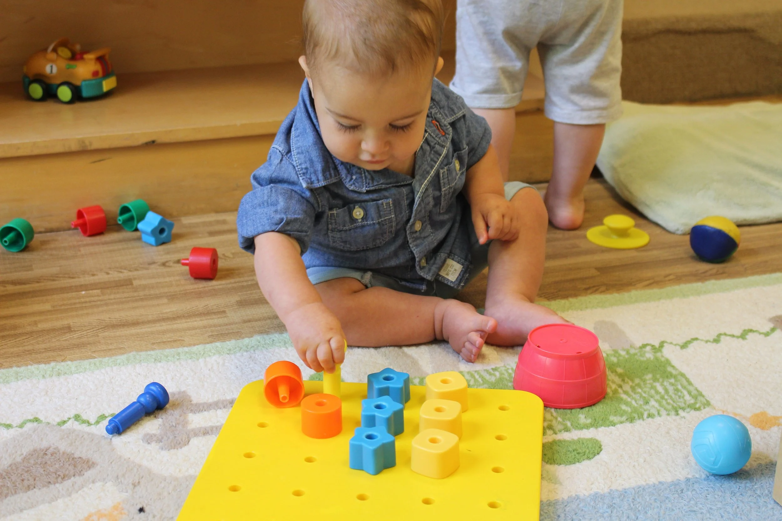 Young child playing with colorful plastic stacking toys on a yellow pegboard in a nursery or playroom.