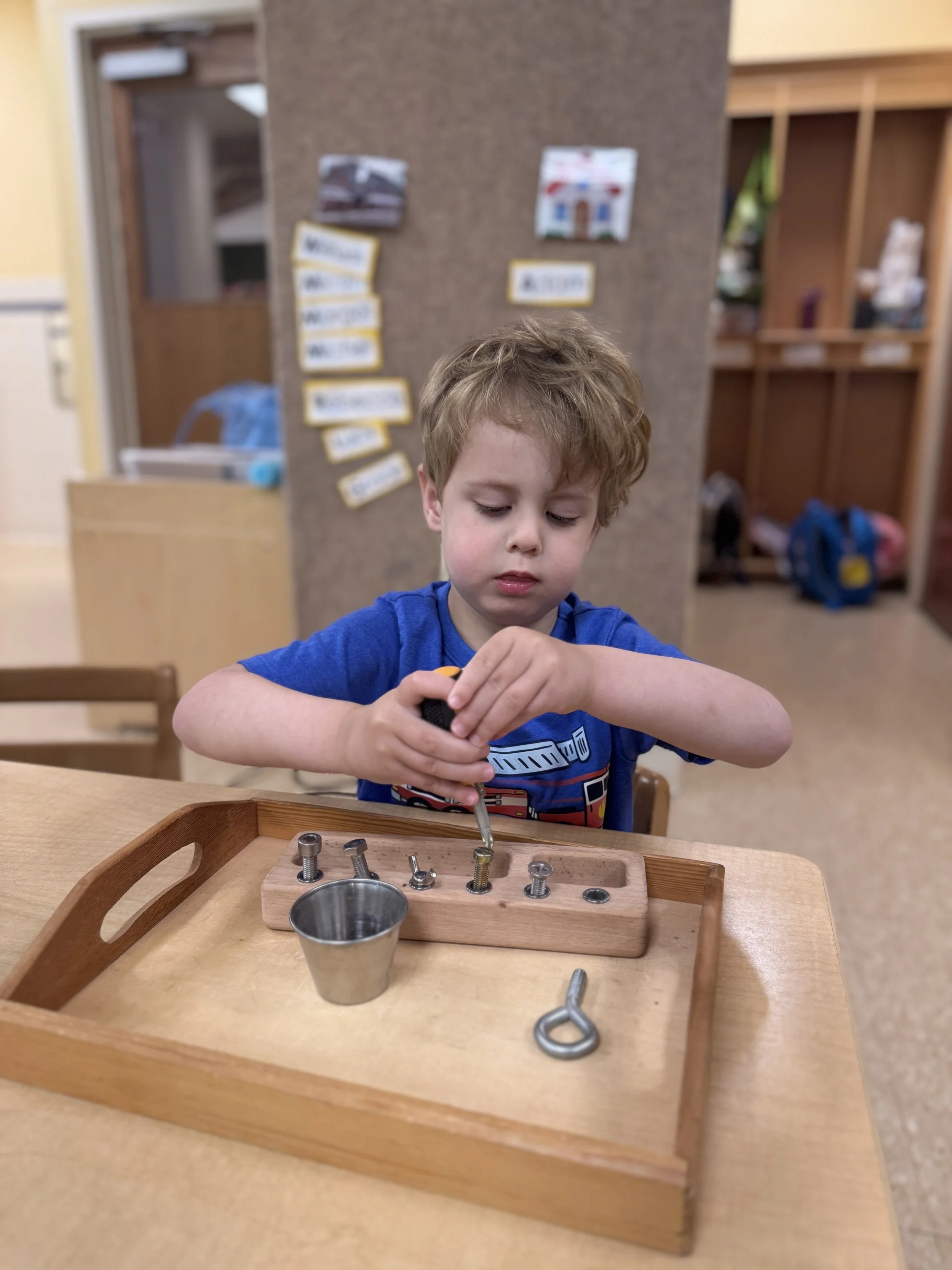 A young boy with curly hair is sitting at a wooden table, working on a woodworking activity with screws and a screwdriver, in a classroom setting.