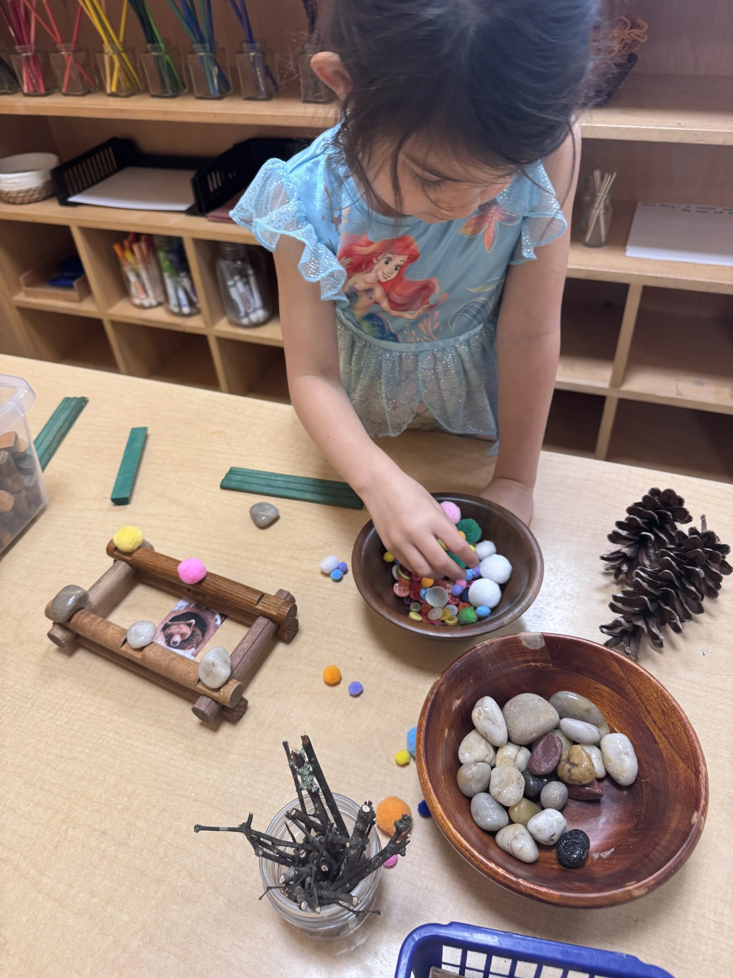 Child wearing a Disney Ariel dress playing with colorful pom-poms, small stones, and craft supplies on a wooden table in a craft room.