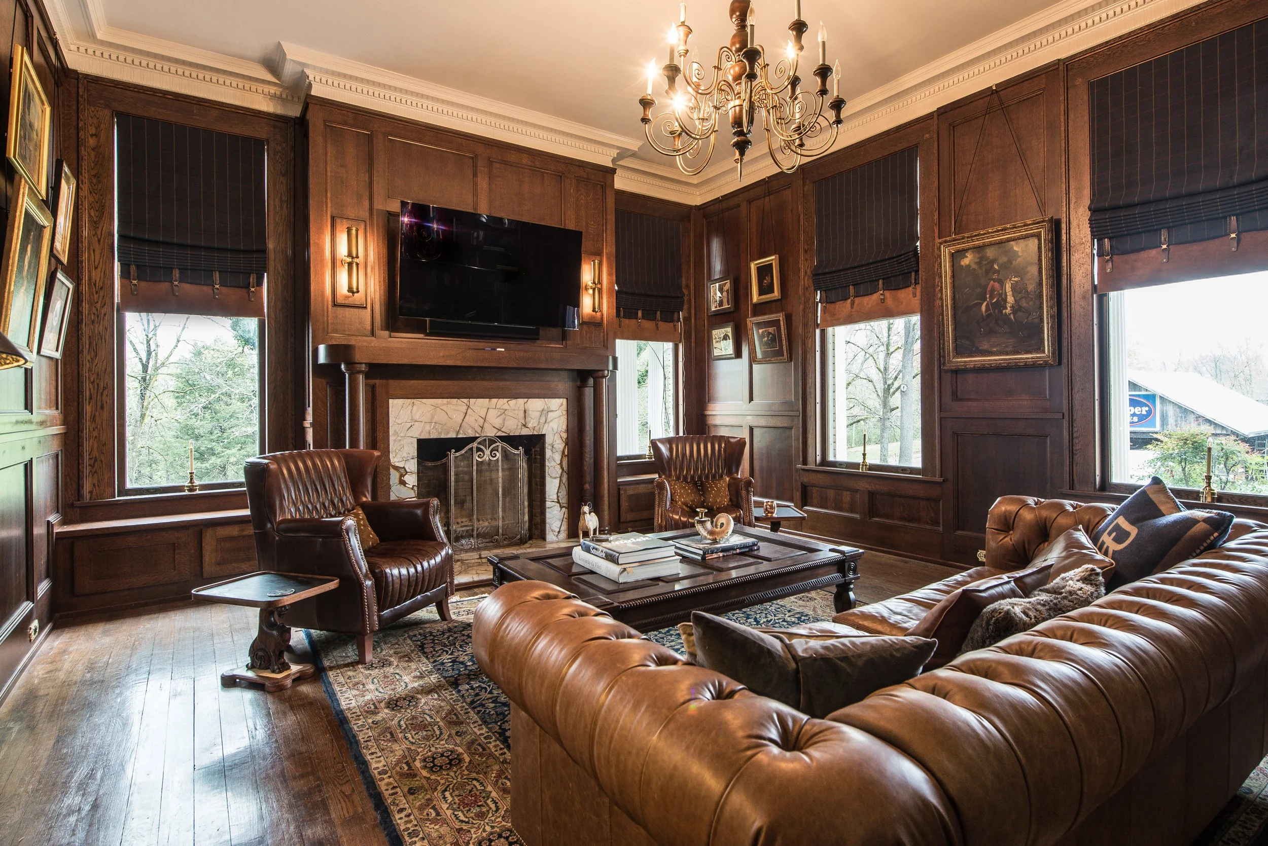 Living room with dark wooden walls, a brown leather Chesterfield sofa, two leather armchairs, a large wooden coffee table, a stone fireplace with a TV mounted above it, window seats with dark shades, and a chandelier hanging from the ceiling.