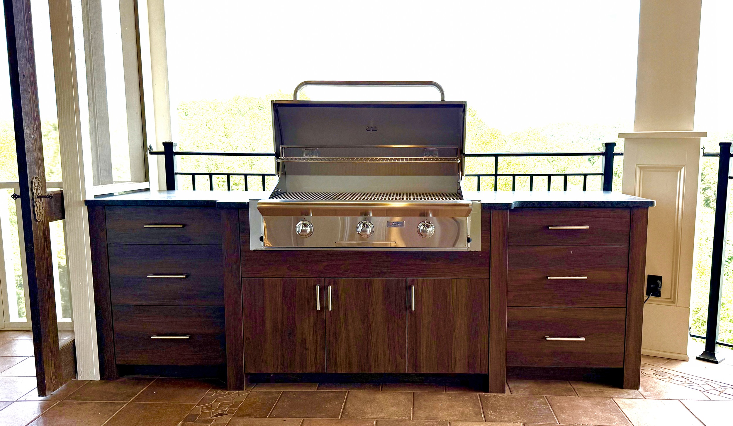 Grill station with a stainless steel barbecue grill placed on a wooden countertop with drawers and cabinets, set on a tiled patio area.