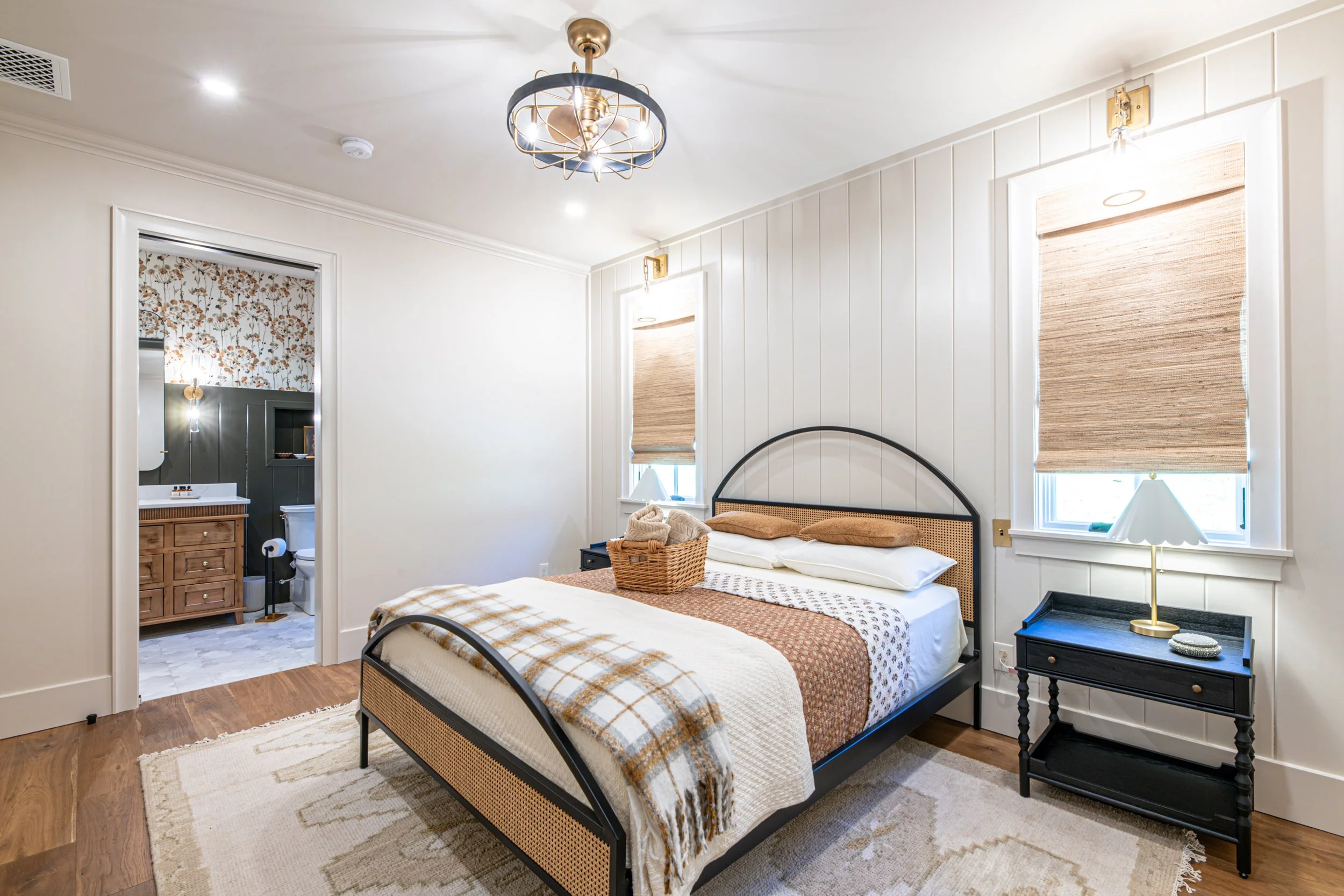 A cozy bedroom with a bed, woven basket, black nightstand with a lamp, two windows with woven shades, and a doorway leading to a bathroom.