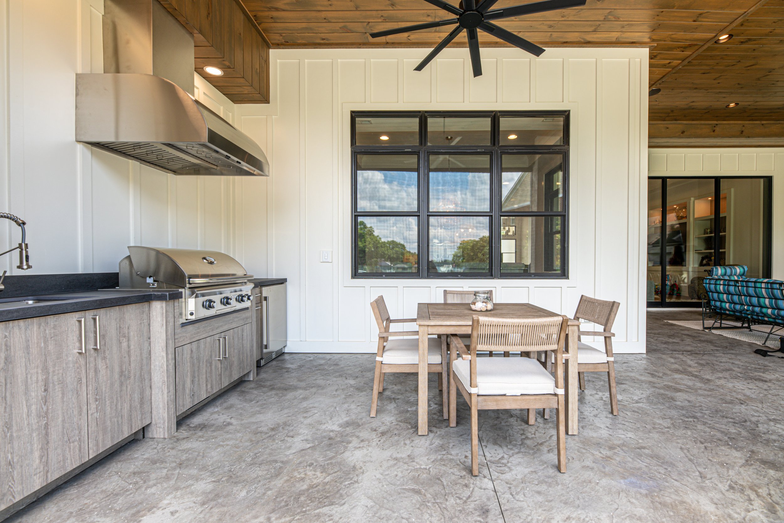 Outdoor kitchen area with a stainless steel grill, wooden cabinetry, and a dining table with four chairs under a wooden ceiling and a large window overlooking a bright outdoors.