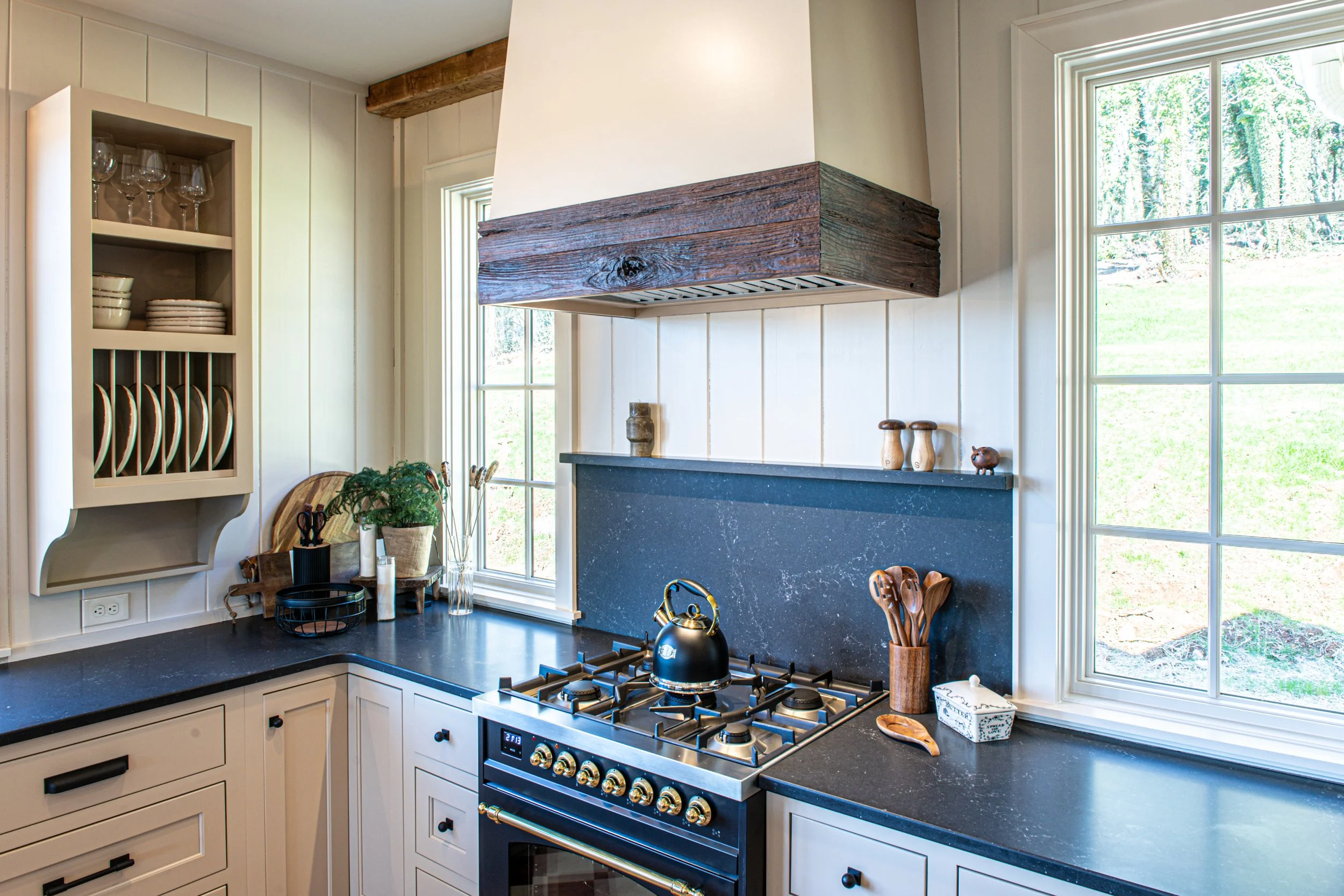 Kitchen corner with black countertop, white cabinets, a stove with a kettle, a window, and decorative items including a plant, wooden utensils, and a tissue box.