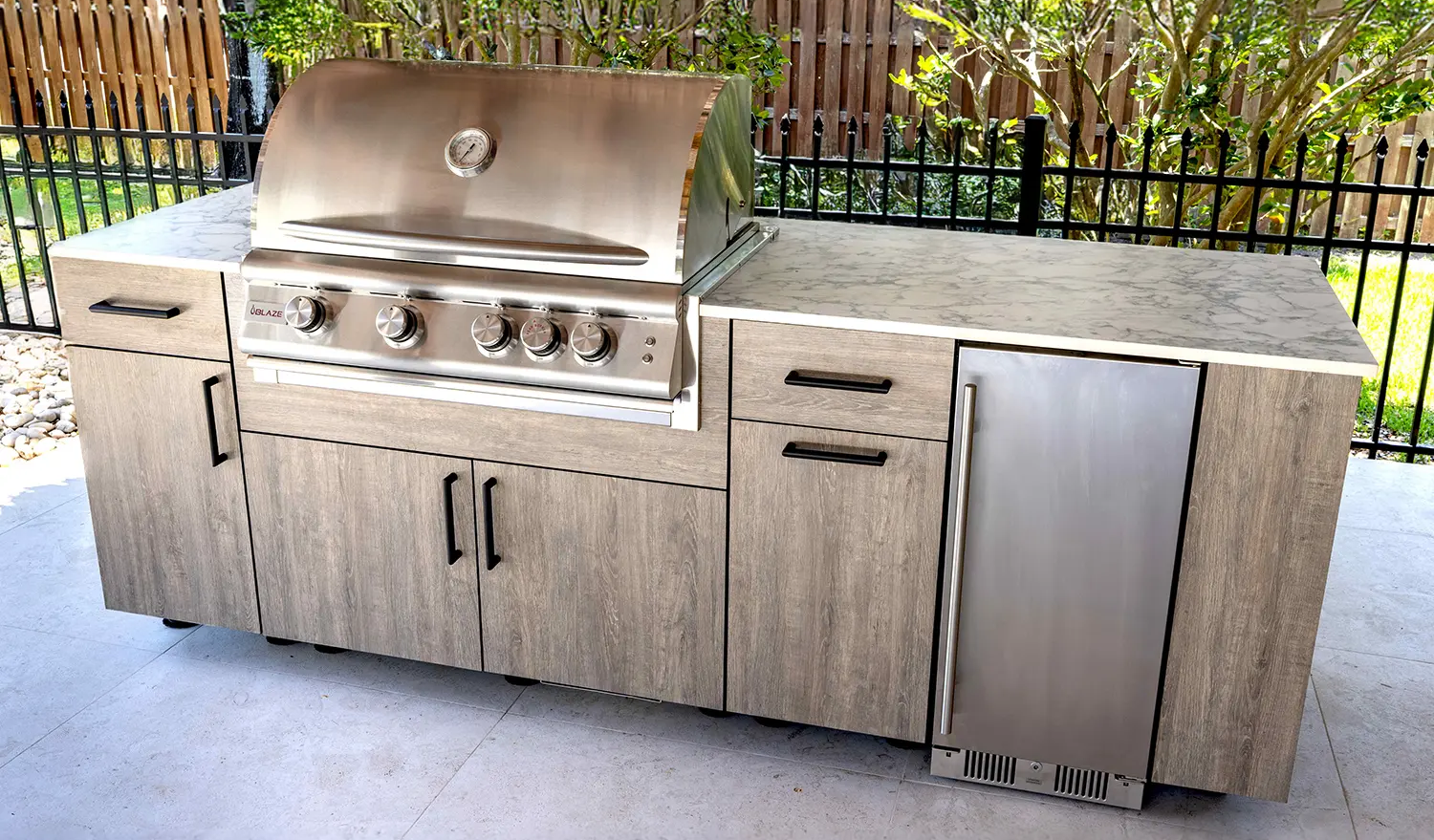 Outdoor kitchen with a stainless steel grill, cabinets, and a marble countertop, surrounded by a wooden fence and greenery.