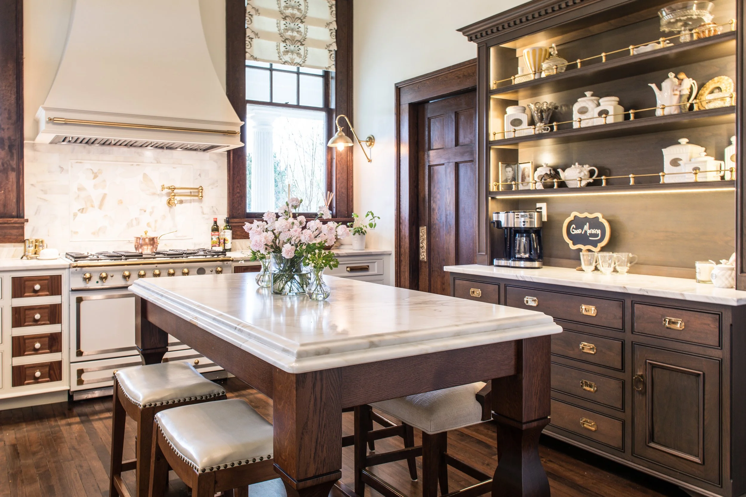 A kitchen with white and dark wood cabinetry, a marble countertop, a kitchen island with a marble top, a flower vase, wall-mounted brass kitchen faucet, open shelves with dishware, a coffee maker, and a window with a roman shade.