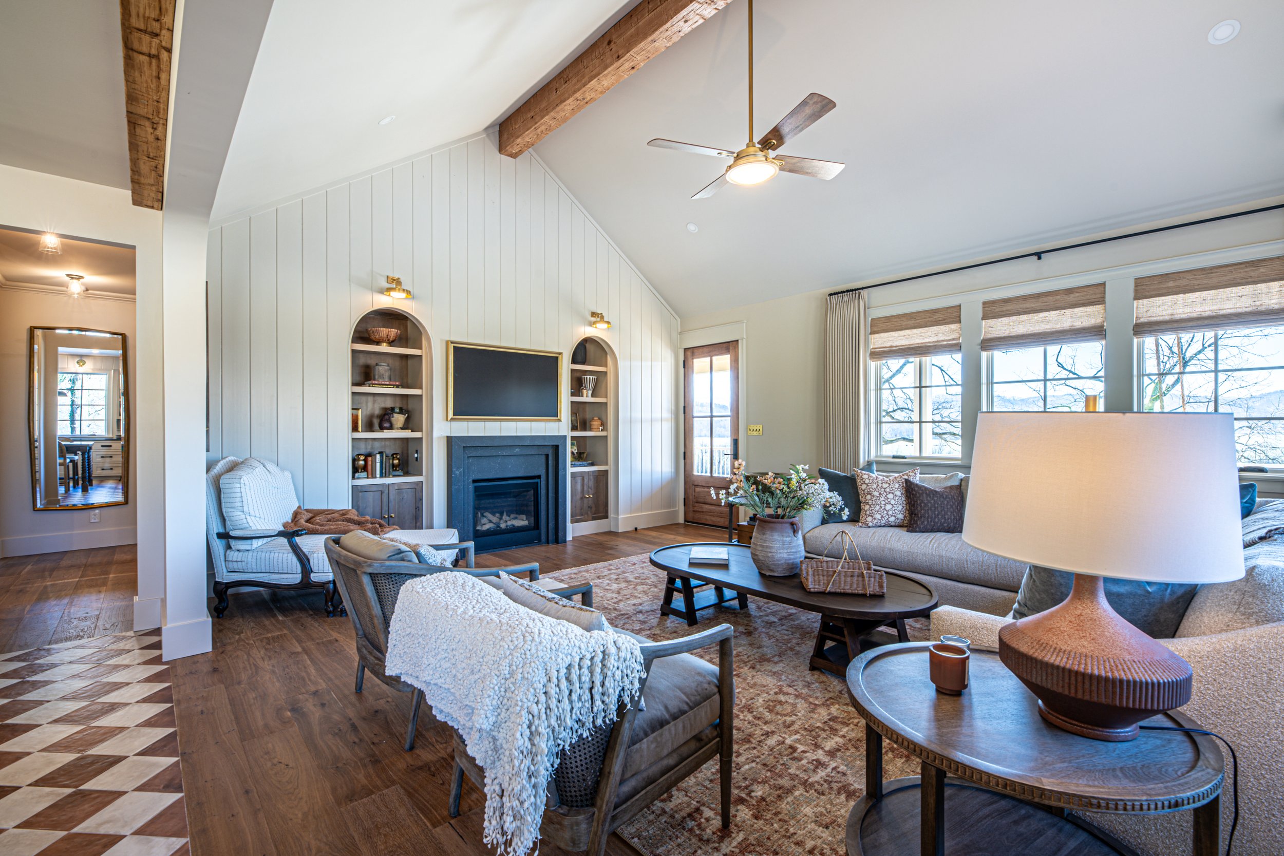 Living room with wood floors, a fireplace with shelves on each side, a large window with blinds, beige sofas, chairs, a coffee table, and a lamp.