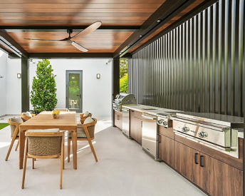 Outdoor patio with wooden dining table, six chairs, and stainless steel barbecue grills, enclosed by a black slatted wall and a white building with a door and greenery.