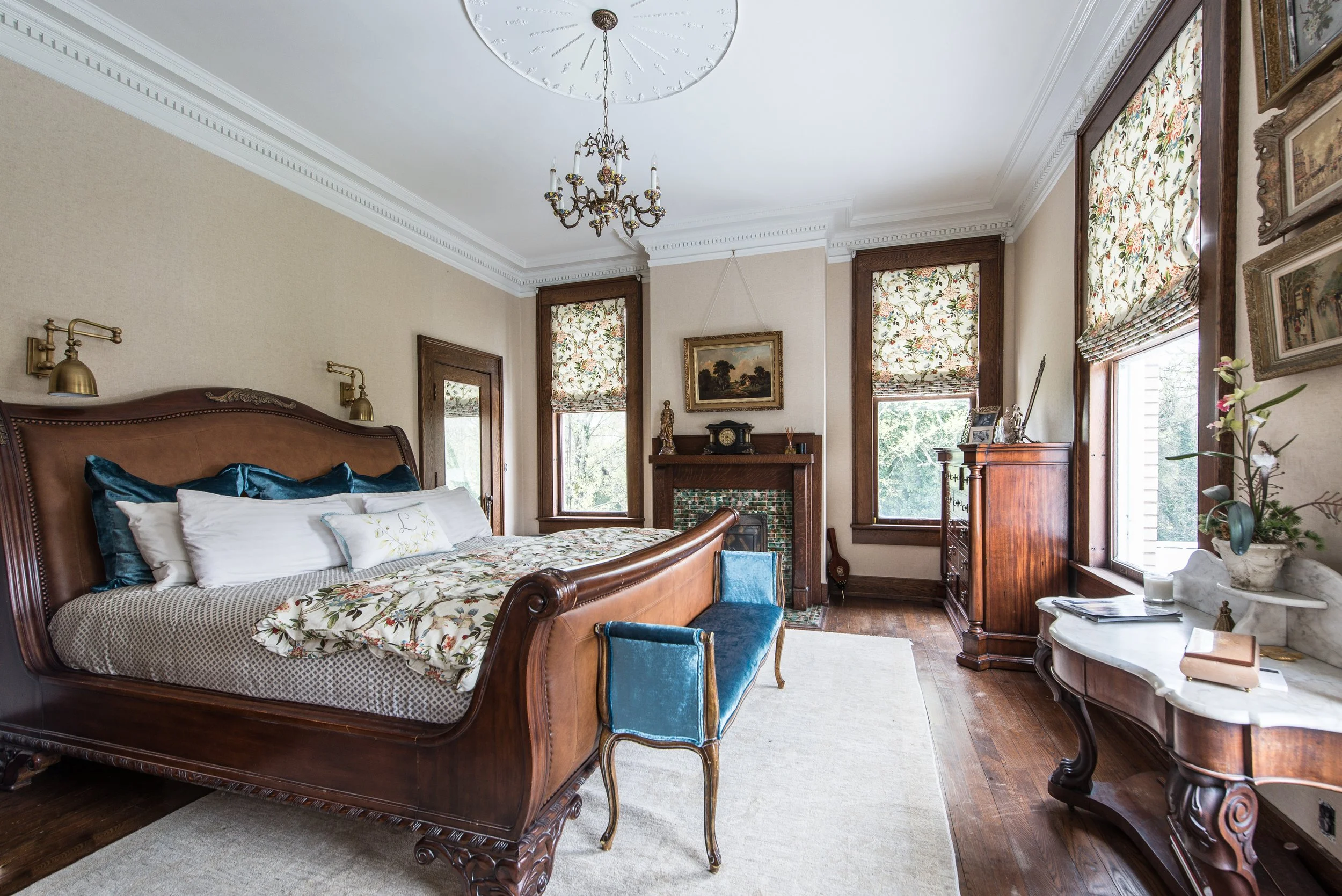 Vintage bedroom with wooden bed, floral bedding, blue velvet bench at foot, fireplace with painting above, large windows with floral Roman shades, marble-topped dresser, chandelier, and decorative items.