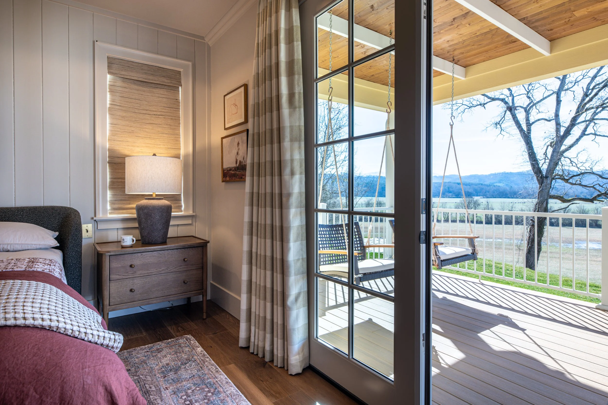 Interior bedroom scene with a bed, side table, table lamp, framed pictures, and sliding glass door leading to a balcony with outdoor swings and a scenic view of trees and mountains in the distance.