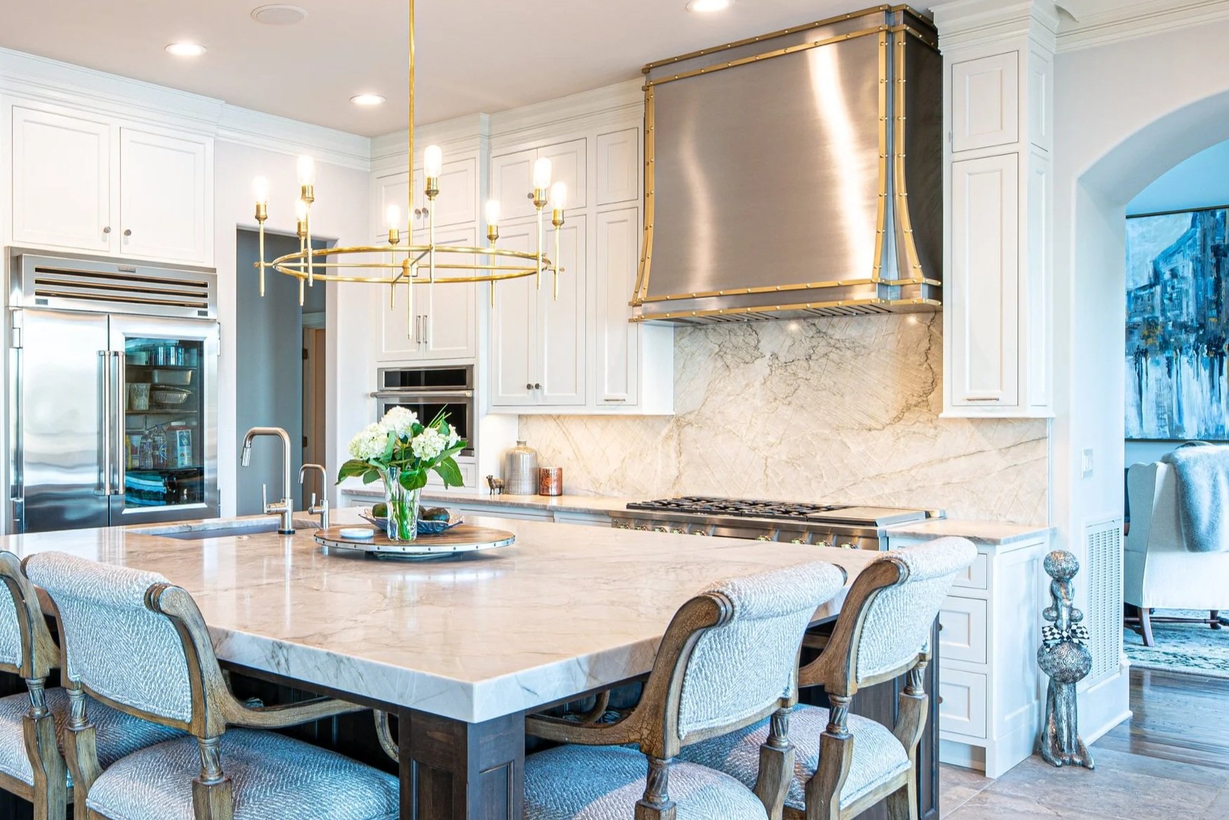 A modern, bright kitchen with white cabinets, a large marble island, a gold chandelier, and stainless steel appliances.