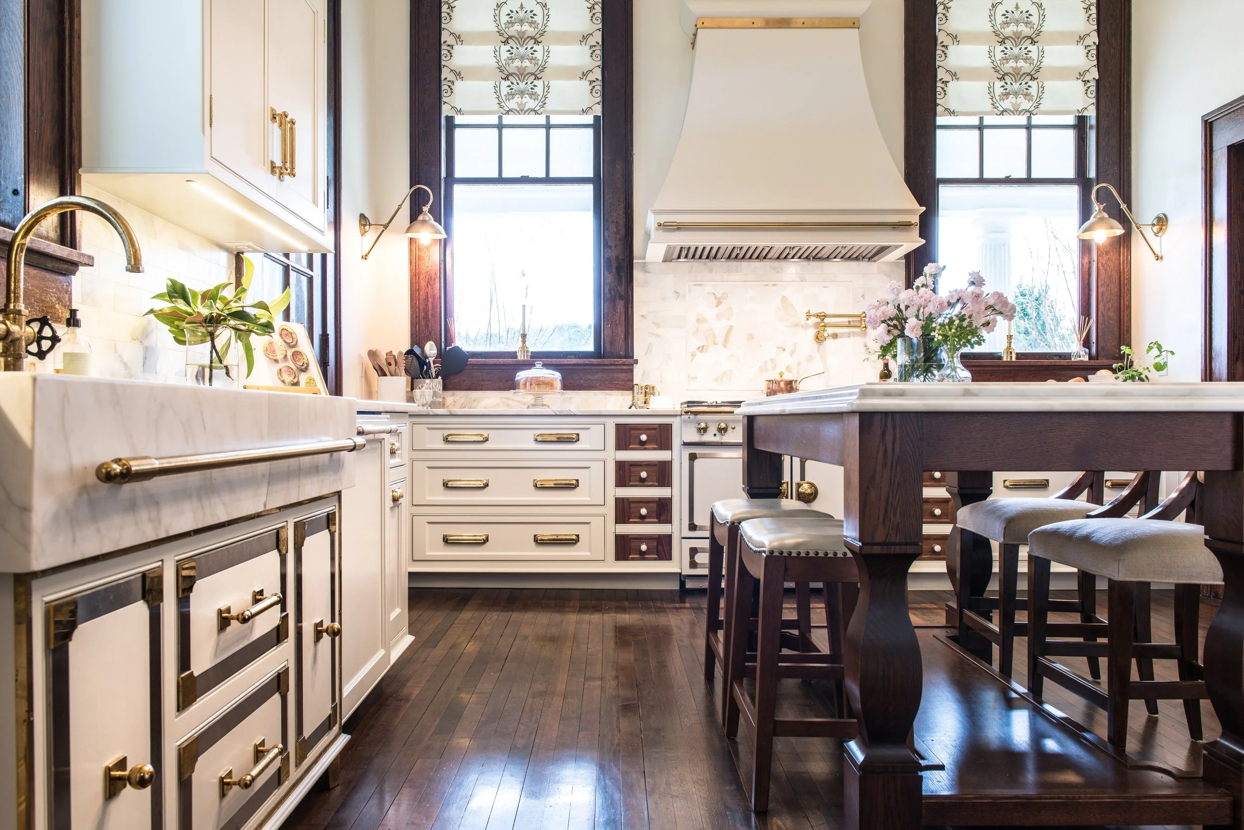 A spacious kitchen with white cabinetry, dark wood accents, and a corner farmhouse sink. Large windows let in natural light, and there are decorative flowers on the counter. The kitchen has a central wooden island with cushioned stools and brass hardware details.