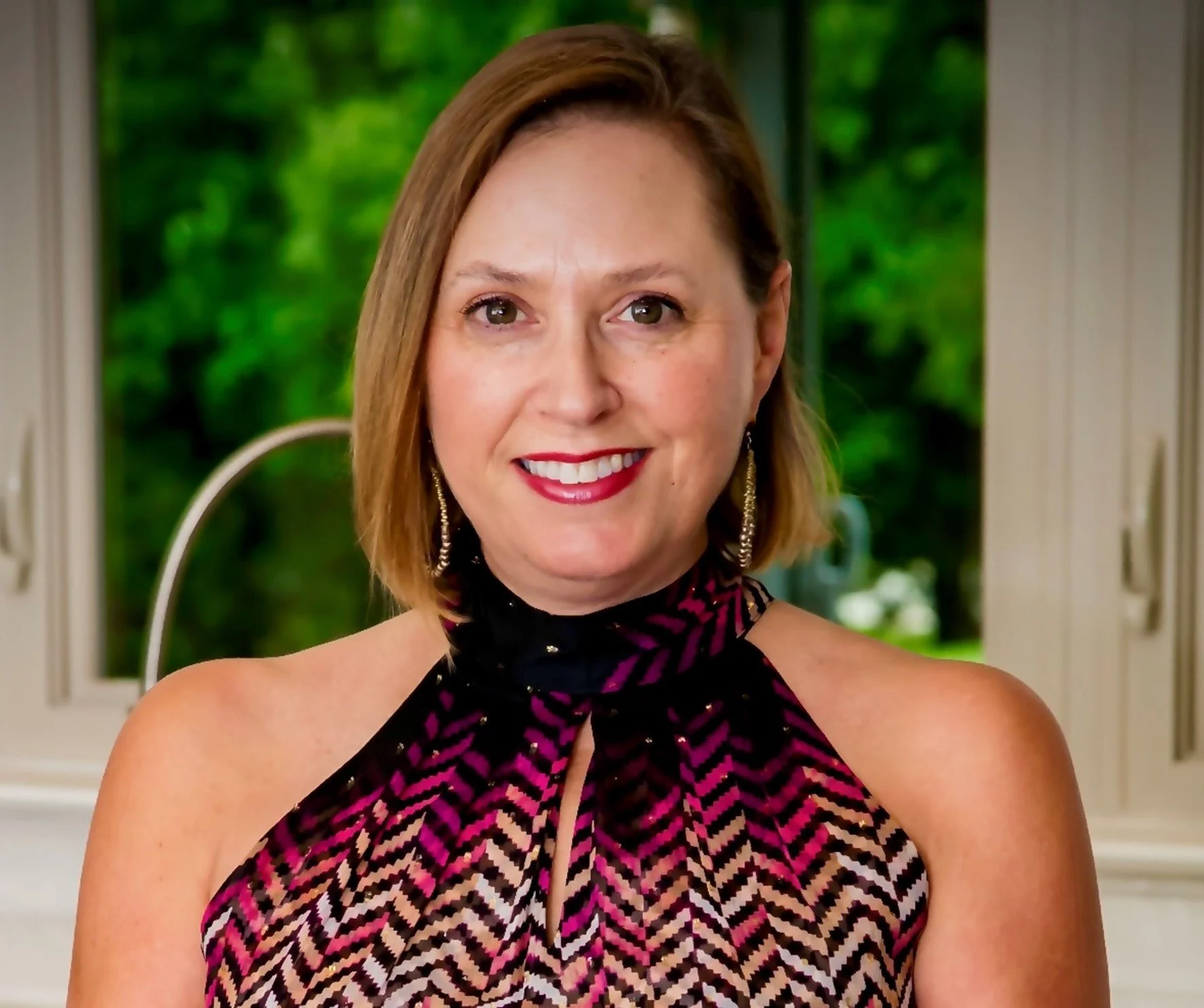 Portrait of a smiling woman with short brown hair, wearing a sleeveless, patterned top, and earrings, standing indoors near a window with greenery outside.