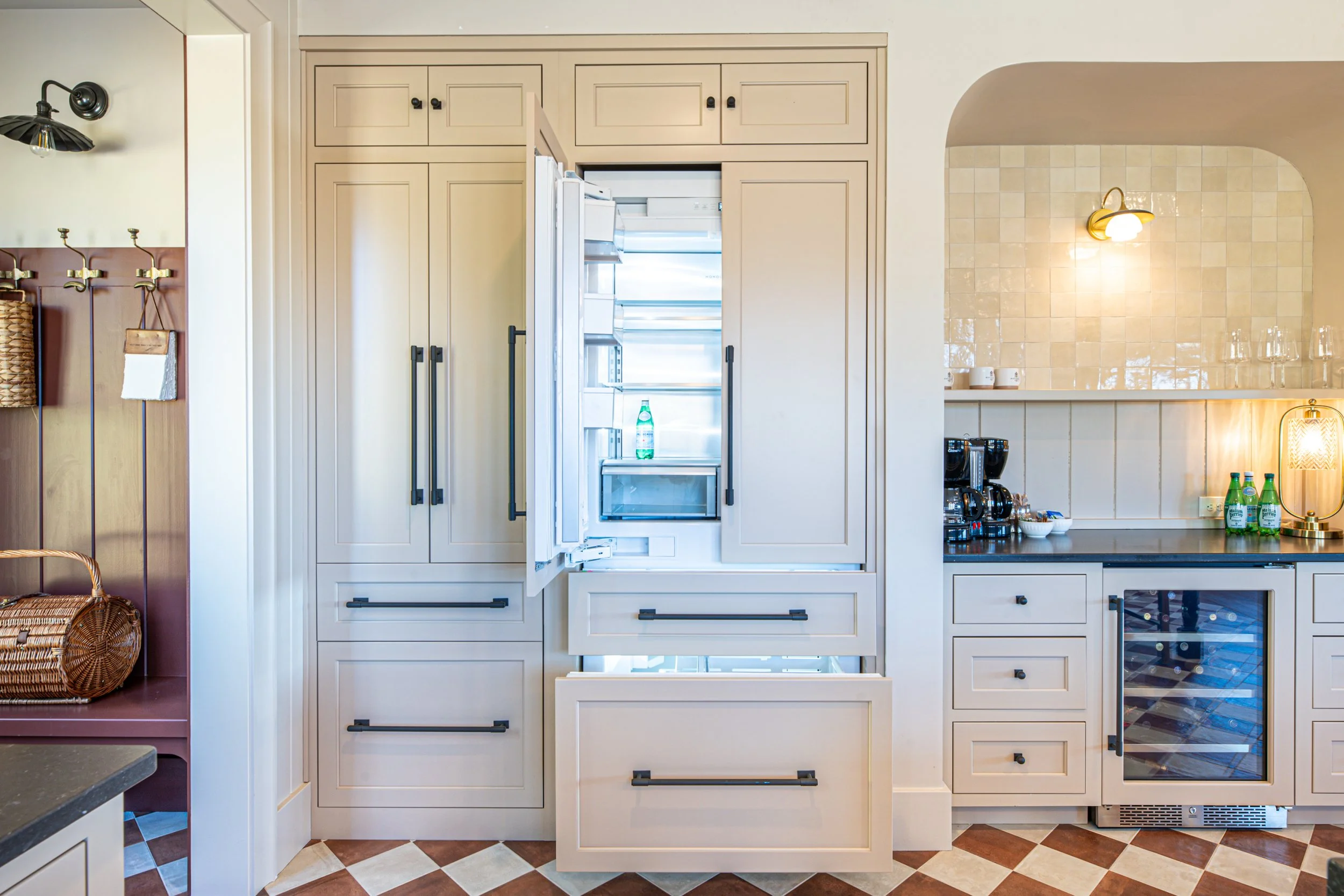 Open refrigerator in a modern kitchen with beige cabinets, black handles, and a wine cooler, next to a coffee station with glasses and bottles of water