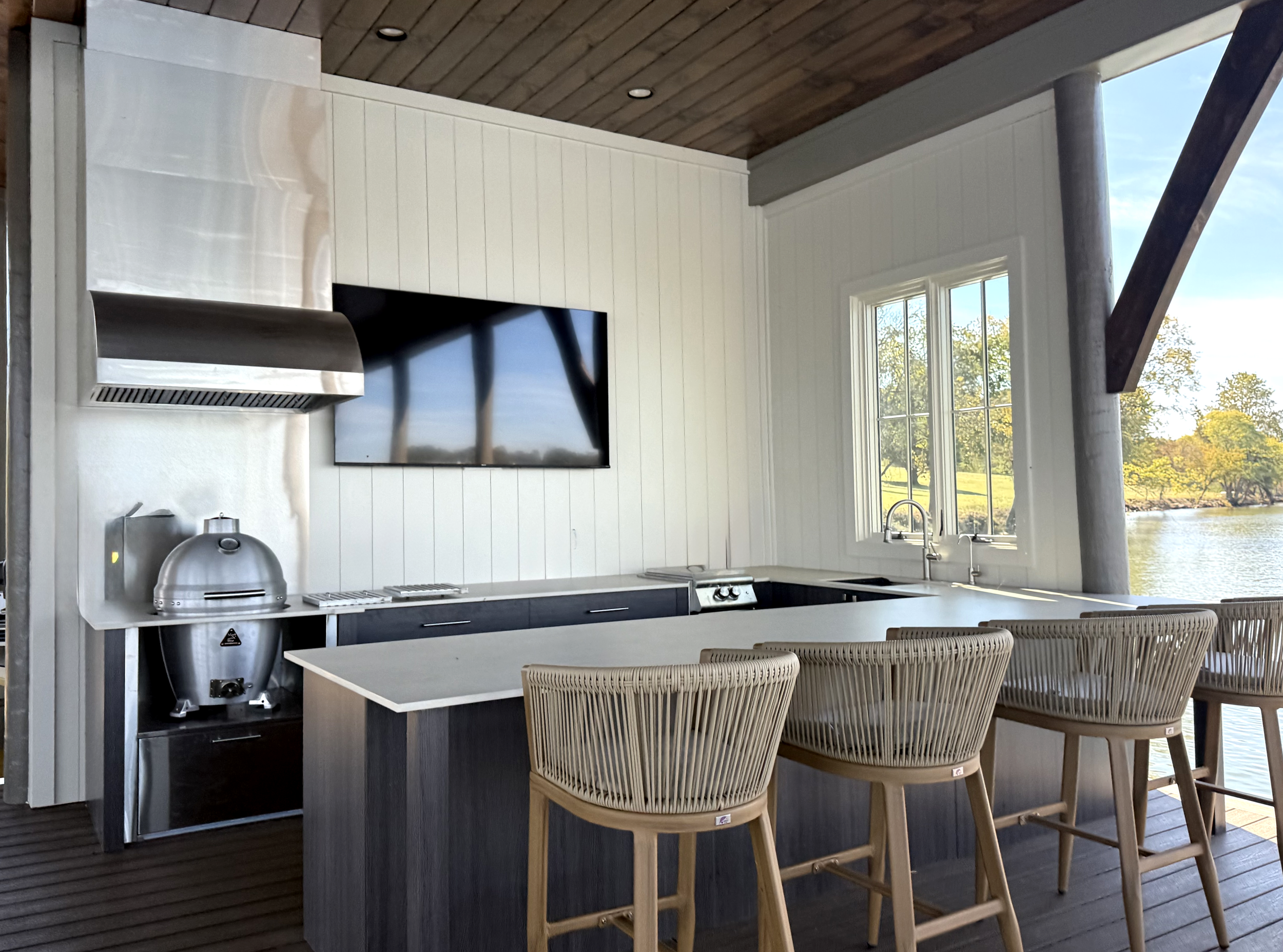 Modern outdoor kitchen with a light-colored countertop, four wicker bar stools, a large window overlooking a river and trees, and a flat-screen TV on a white wood-paneled wall.