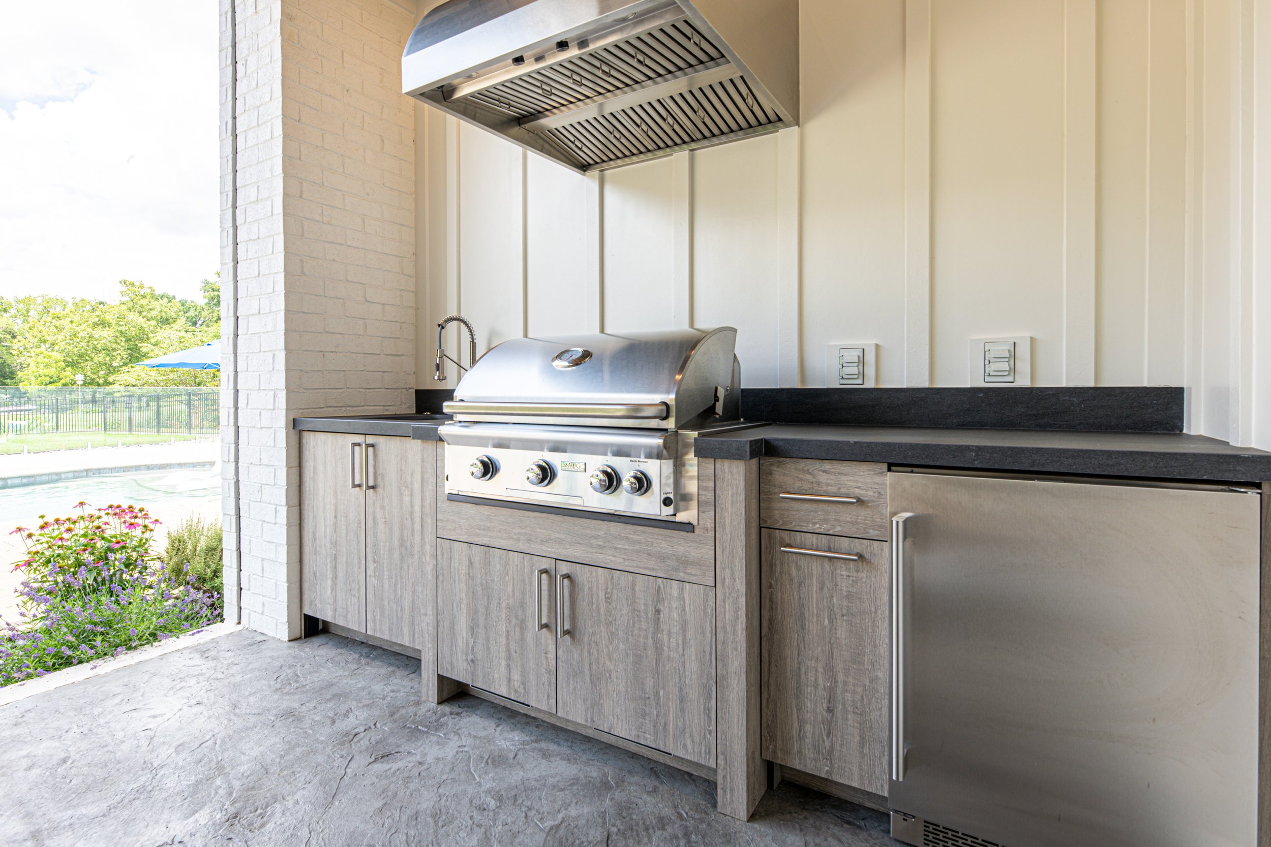 Outdoor kitchen area with a stainless steel gas grill, countertop, and cabinetry, with a pool and trees visible in the background.