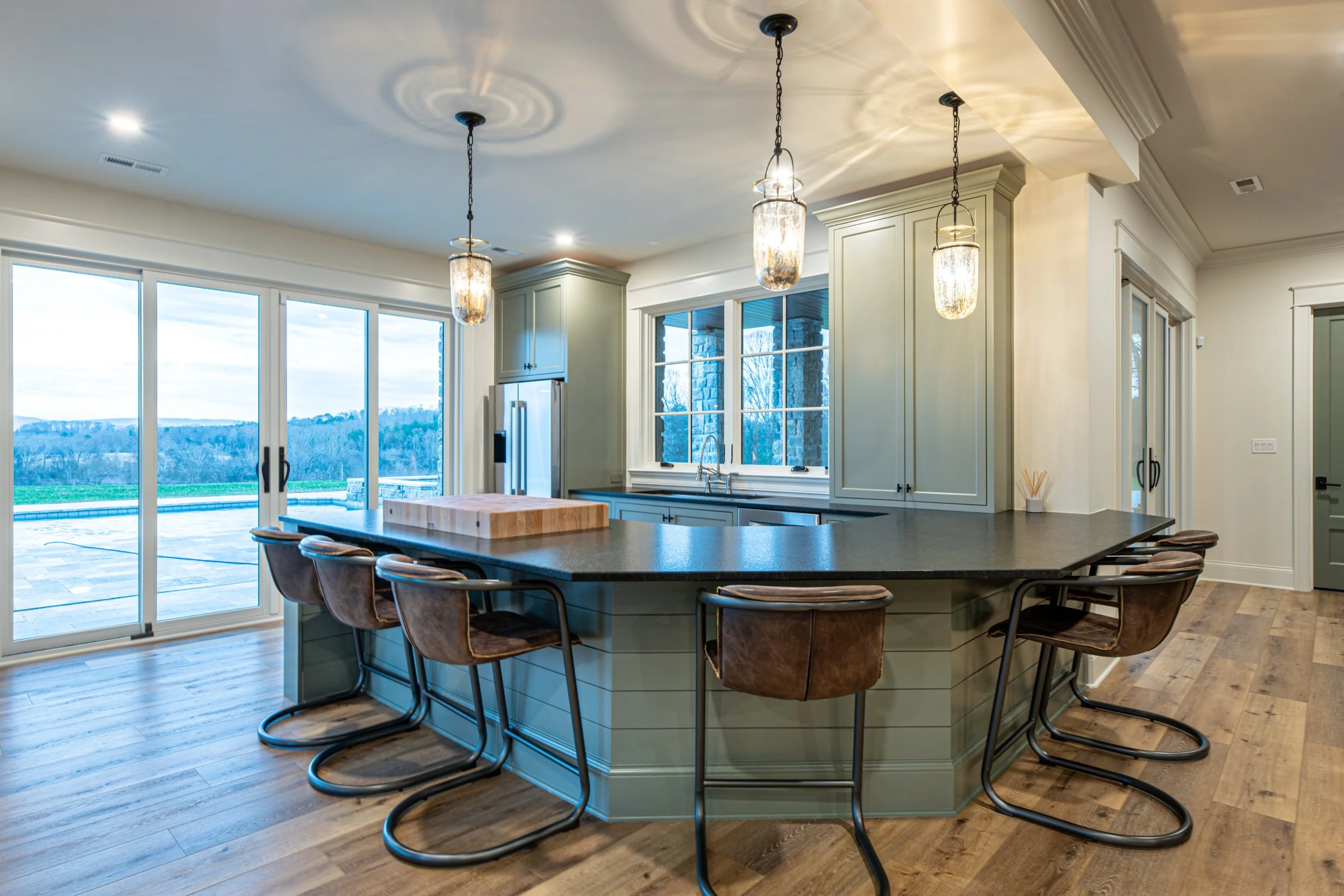 Modern kitchen with green cabinetry, black countertop, central island with bar stools, large sliding glass door, and pendant lights.