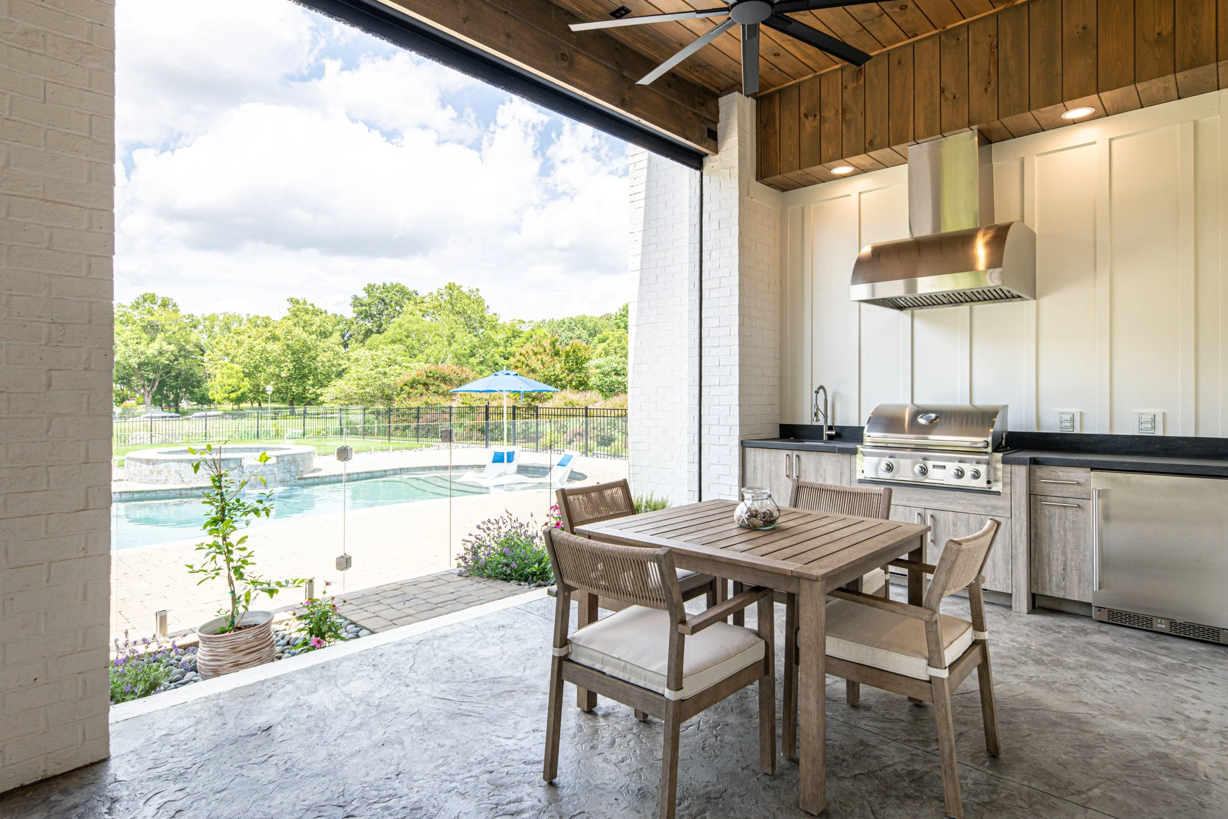 Outdoor patio area with a dining table and chairs, kitchen grill, and a view of a backyard with a pool and lounge chairs under an umbrella.