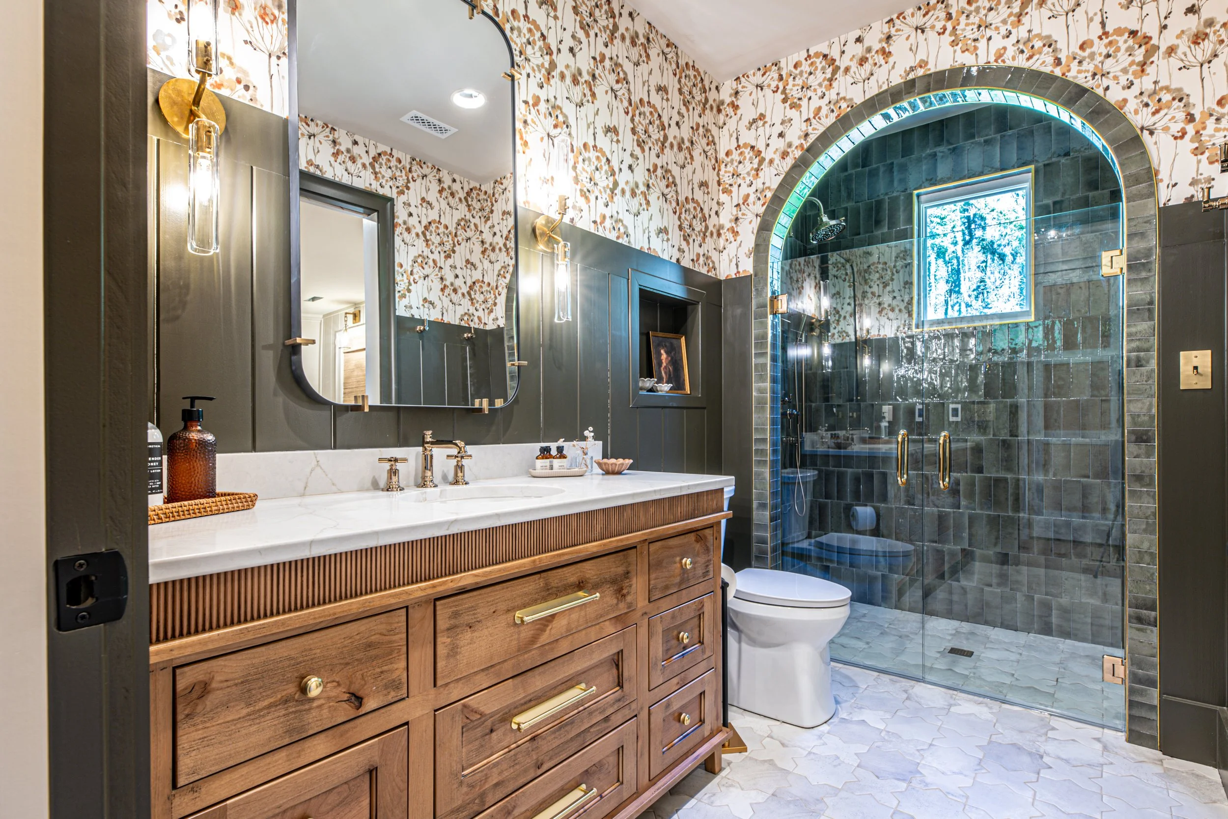 Modern bathroom with floral wallpaper, wooden vanity with marble countertop, and glass-enclosed shower with navy blue tiles and a window.