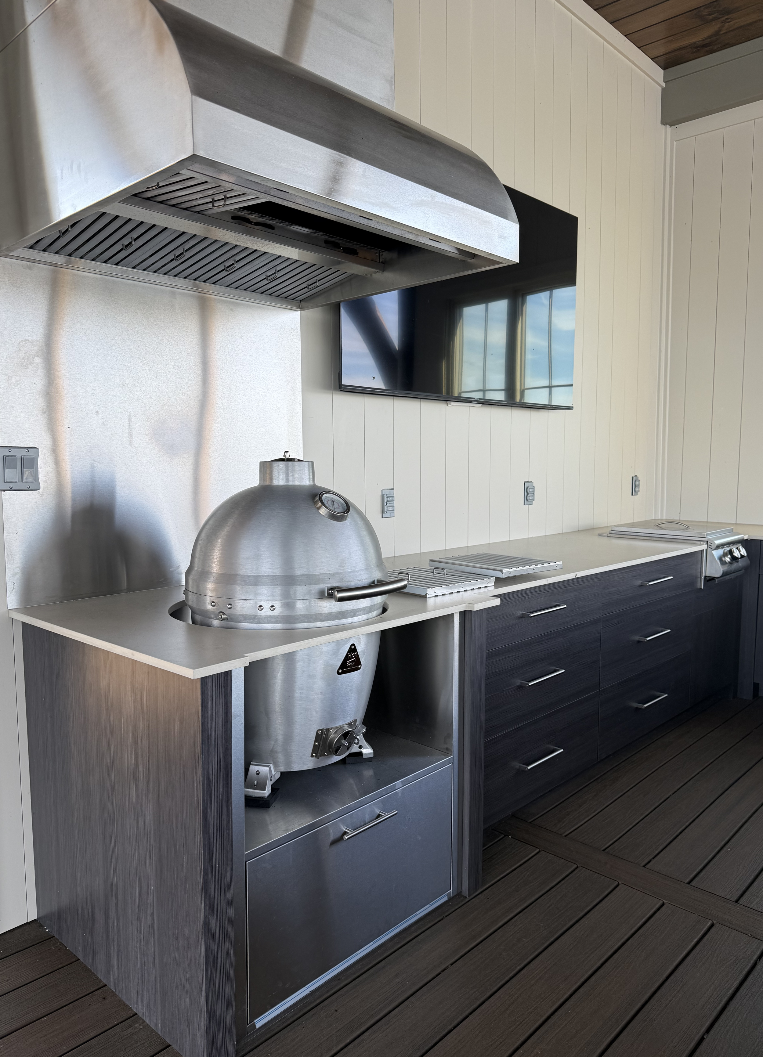 Outdoor kitchen area with stainless steel grill, hood, and a black cabinet with drawers, against white wooden panel walls and a wooden ceiling, on a dark wood deck.