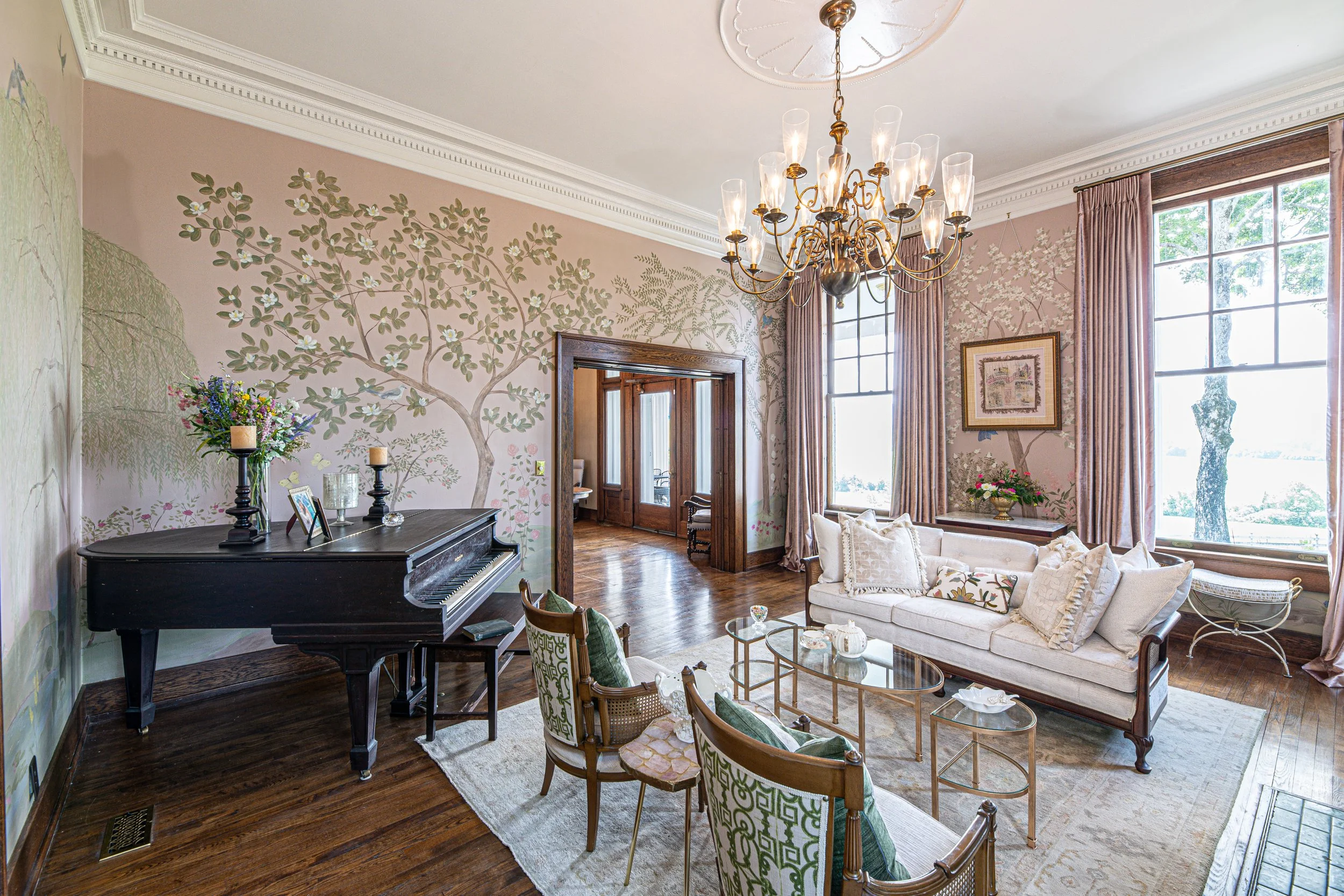 Living room with a grand chandelier, floral wallpaper, a black piano, a cream sofa with decorative pillows, wooden chairs, and large windows with pink curtains.