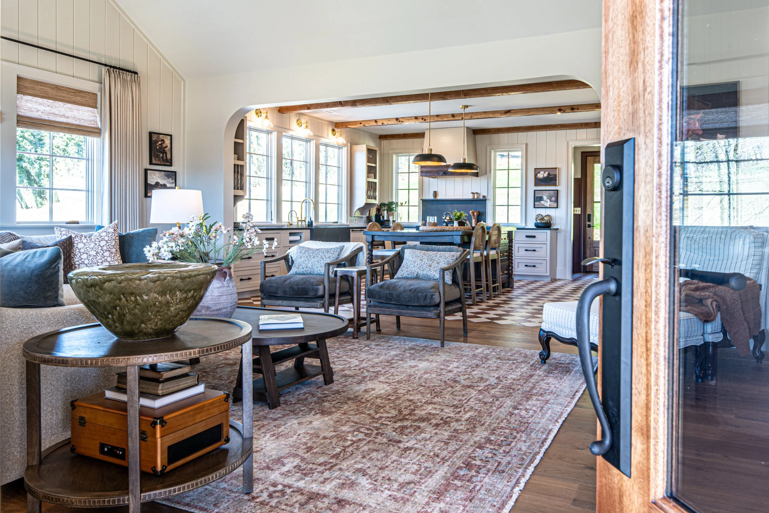 Living room with front door open, view into kitchen with island counter and barstools, windows allowing natural light, and vintage-style furniture including a sofa, armchairs, and a coffee table.