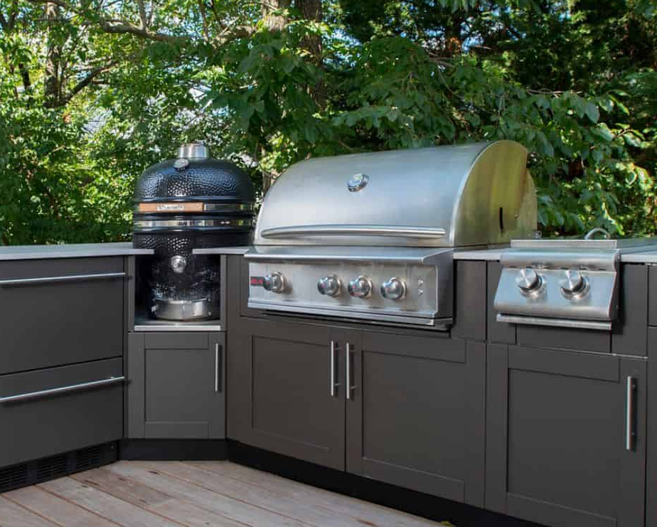 Outdoor kitchen with stainless steel grill, smoker, and storage cabinets, surrounded by green trees and wooden decking.
