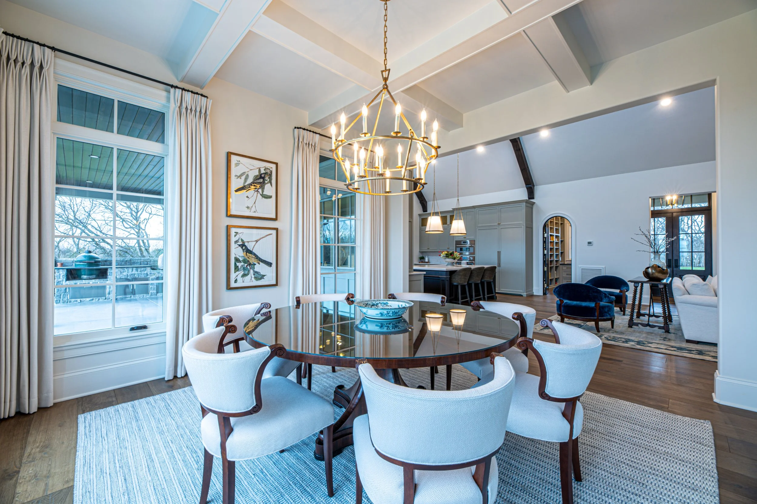 Spacious open-plan dining and living area with a round dining table, upholstered chairs, large windows, chandelier, and a kitchen in the background.