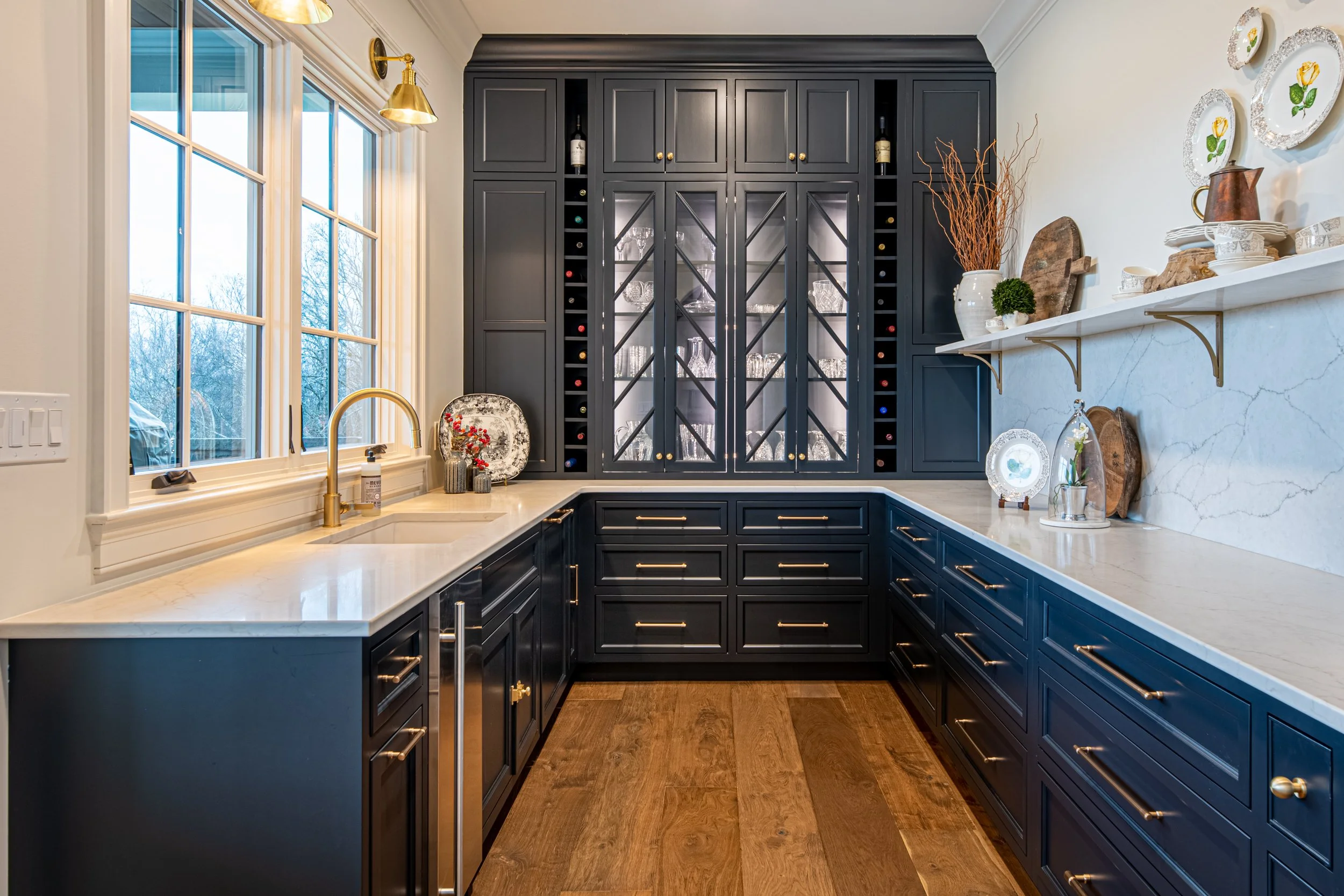 A kitchen with navy blue cabinets, white marble countertops, a large window with a gold faucet, and a built-in wine storage cabinet in the background.