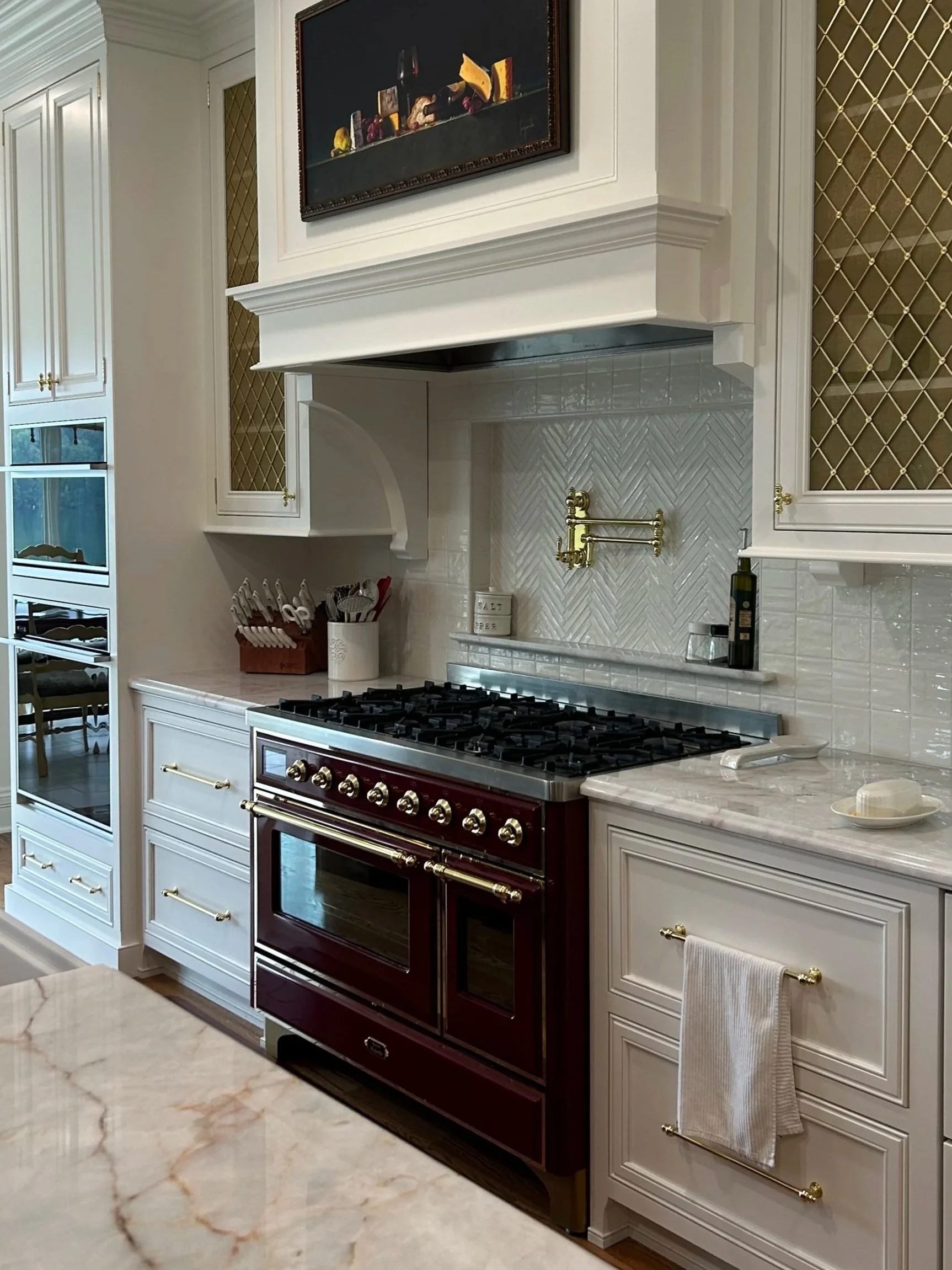 Kitchen with a maroon oven, white cabinets with gold handles, a marble countertop, and a white tiled backsplash.