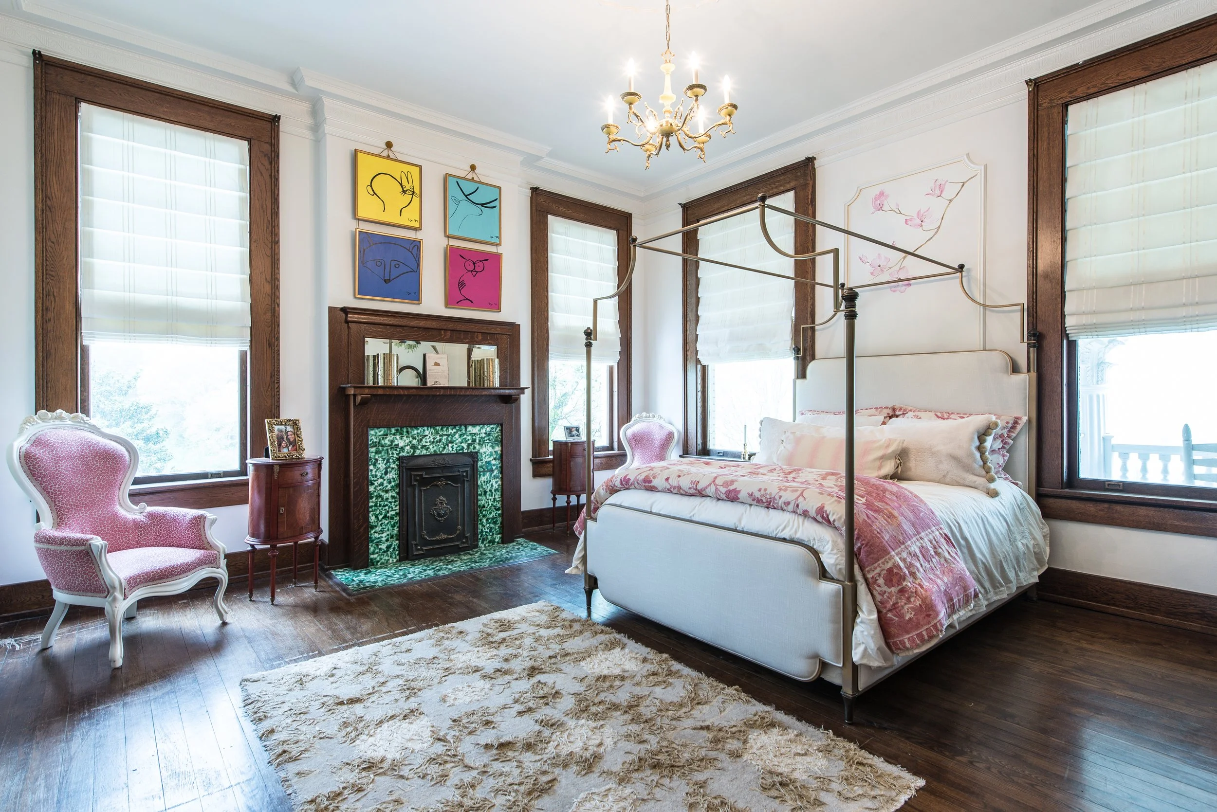 A bedroom with a white bed, pink bedding, four large windows with wooden frames and white shades, a green tiled fireplace, pink armchair, small wooden table, framed photos, colorful artwork with cartoon characters, a chandelier, cream-colored rug, an