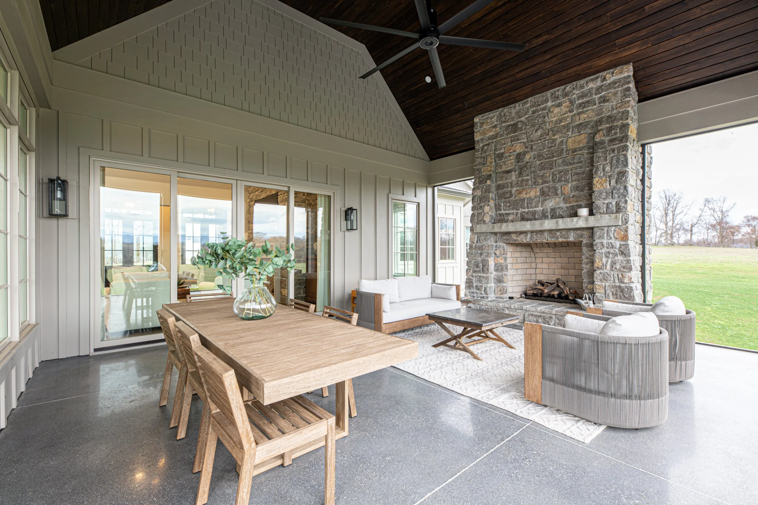 Covered patio with stone fireplace, wooden ceiling, white walls, wooden dining table with chairs, and cozy seating area with sofas and armchairs, overlooking a grassy yard.