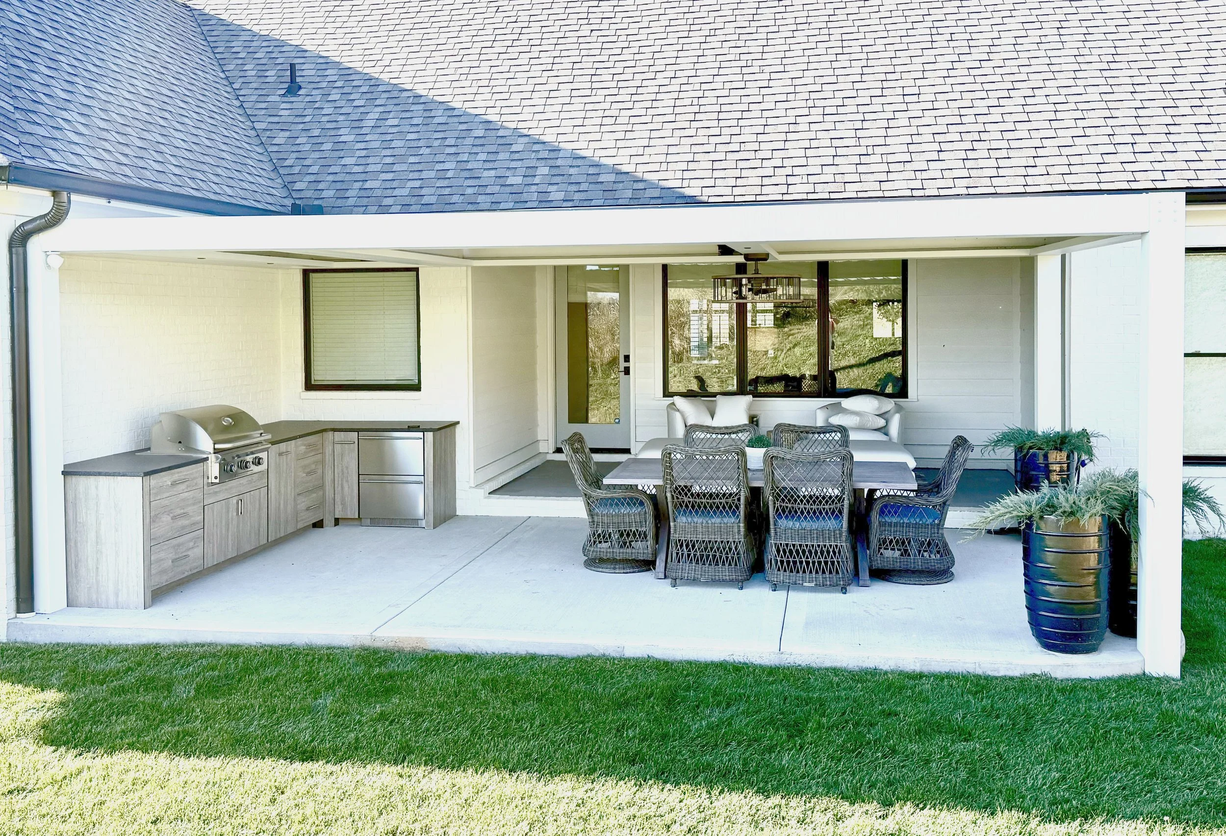 Covered patio with outdoor dining table, wicker chairs, white sofas, potted plants, and outdoor grill, attached to a house with large windows and a shingled roof.