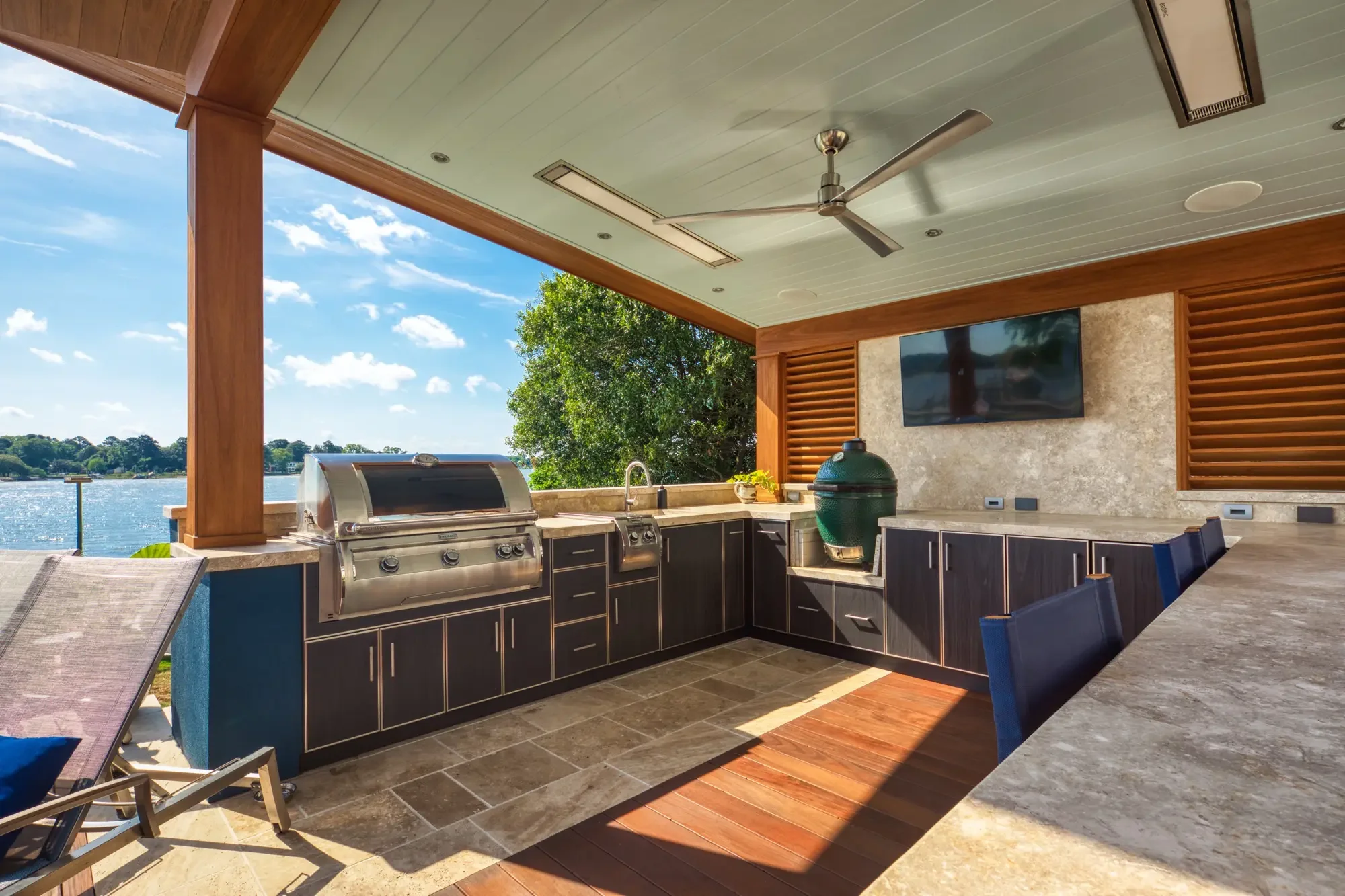 Outdoor kitchen with grill, sink, and TV overlooking a water body with trees and blue sky.