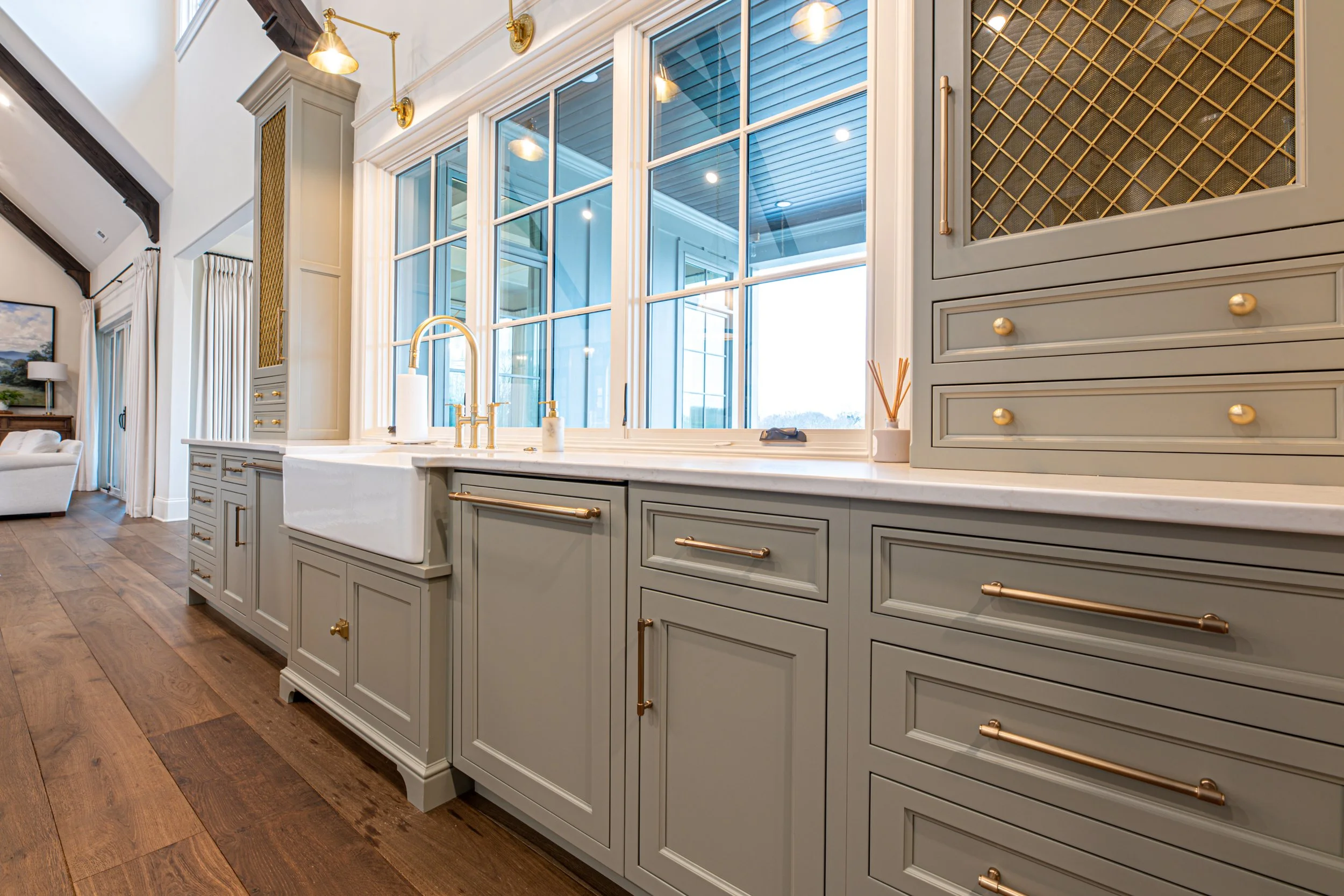 Bright kitchen with gray cabinetry, large window, white farmhouse sink, gold hardware and fixtures, hardwood floors, and view of a sunny outdoor space.