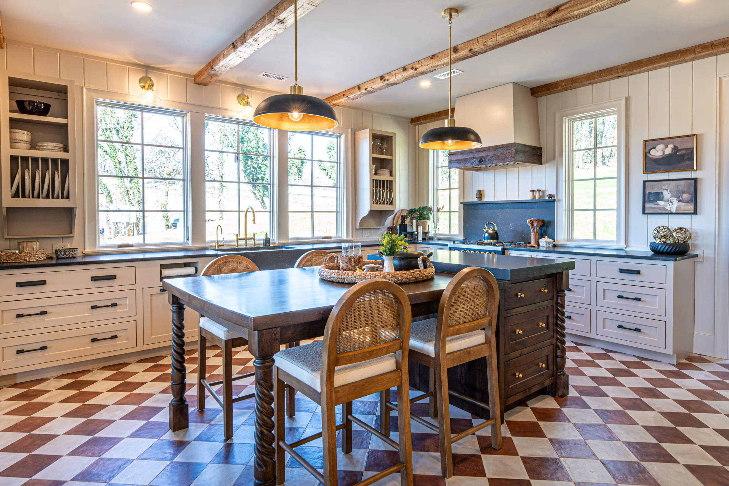 Kitchen with white cabinets, black countertops, and a central wooden island with bar stools. Large windows let in natural light, and there are two black pendant lights hanging above the island. The floor has a checkered pattern with tan and cream til