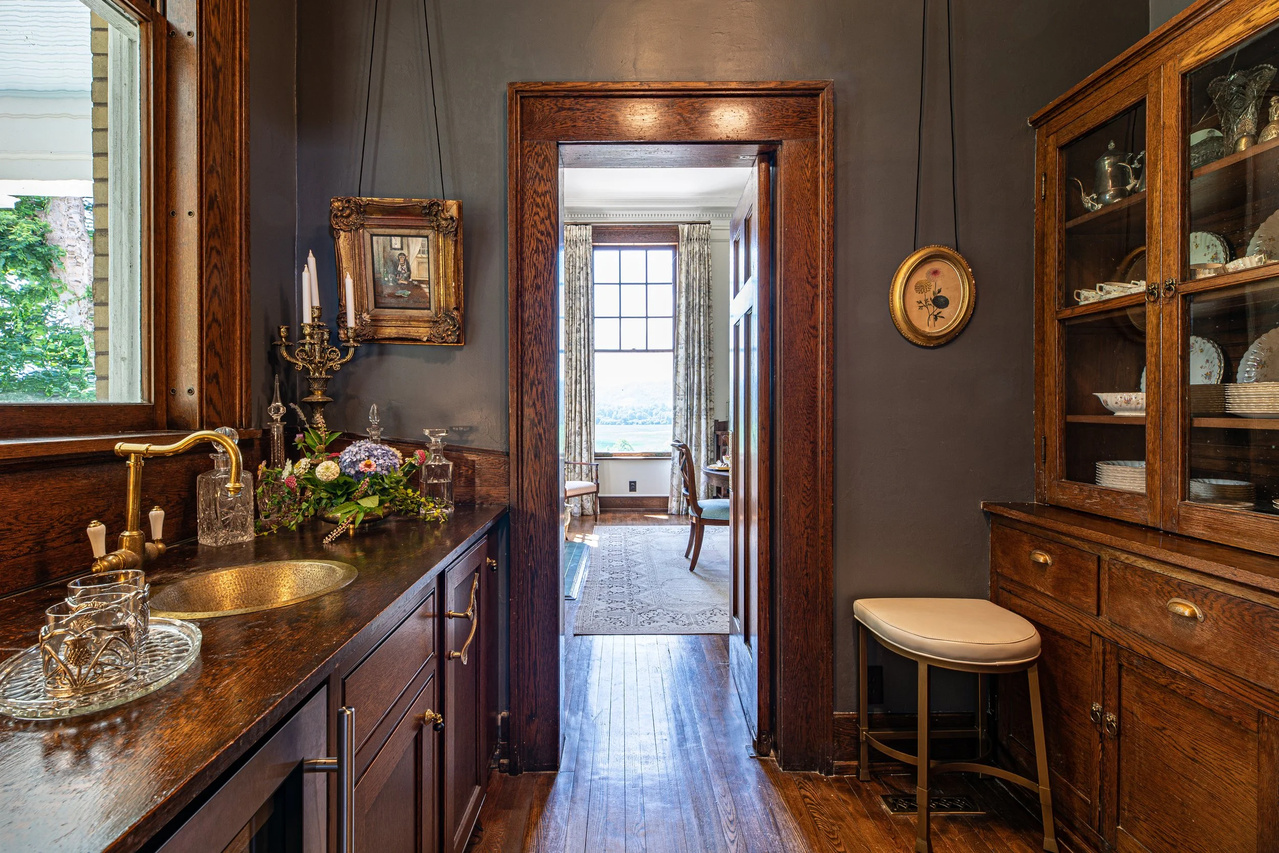 A vintage-style kitchen nook with a dark wood cabinet, a small cream stool, decorative plates, and a window with view of a garden, with a doorway leading to a sunlit dining area with a large window and patterned curtains.