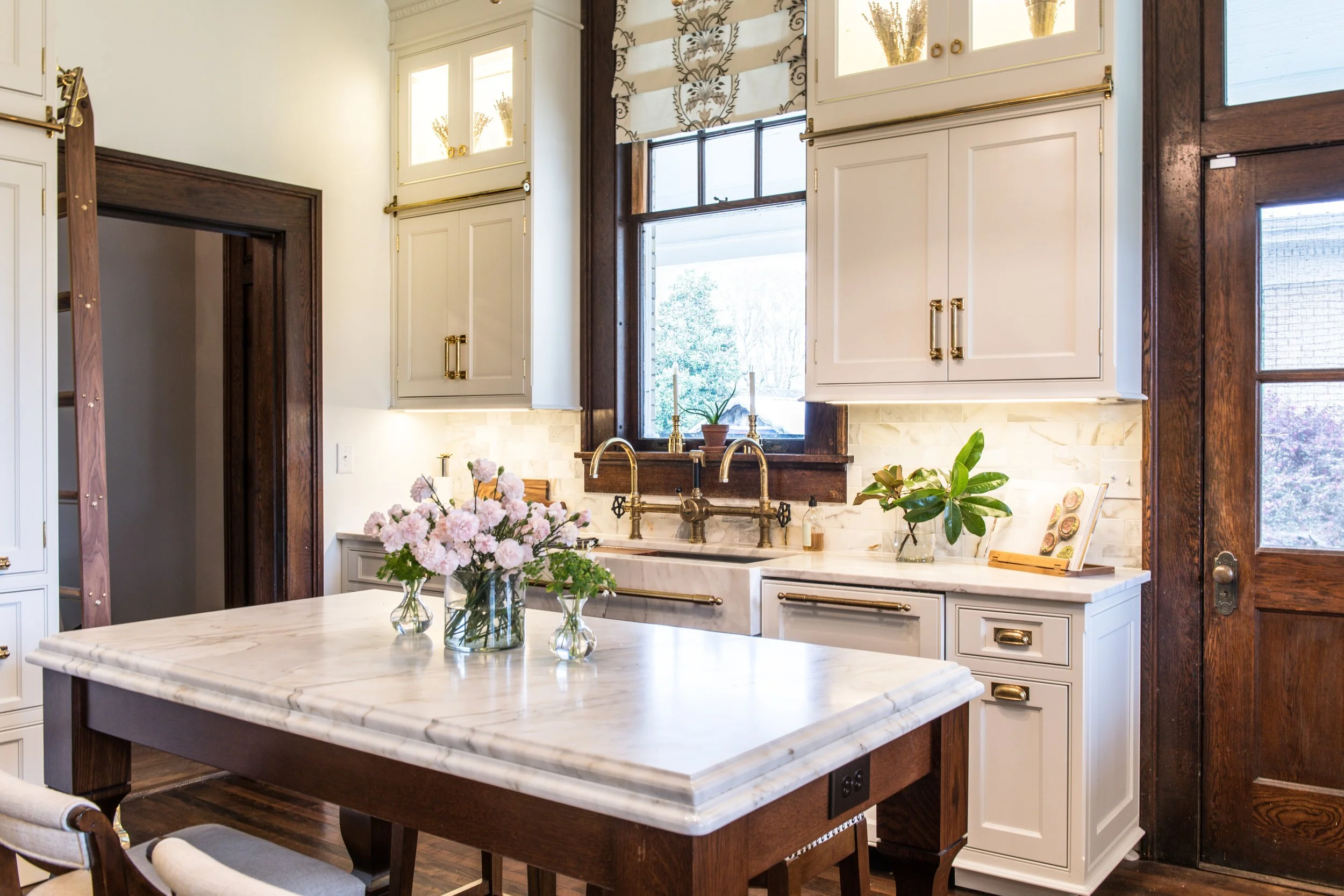 A bright kitchen with white cabinetry, a marble countertop, and a window above the sink with a view of trees outside. The kitchen has plants, flowers, and brass fixtures.