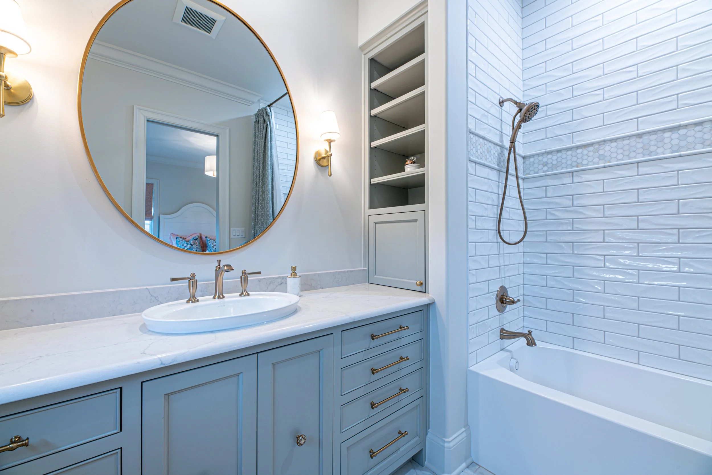 Bathroom with a white vanity sink, a large round mirror, brass fixtures, a built-in cabinet, and a bathtub with a white tiled shower area with a hand-held shower head.