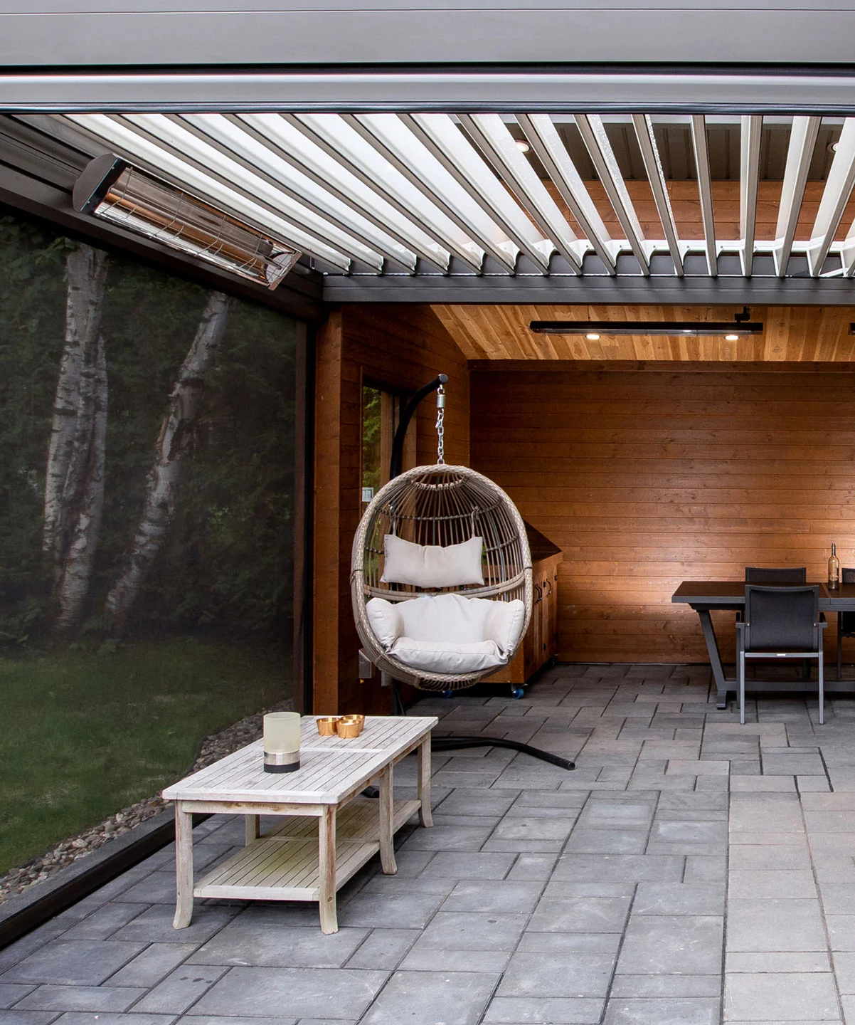 Indoor patio with wooden wall, hanging wicker chair with white cushions, small white table with candles, dining table with chairs, and large glass window showing trees outside.