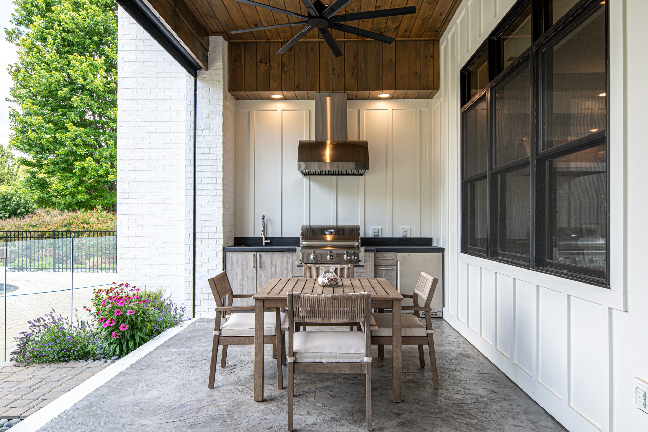 Outdoor kitchen area with a wooden dining table, six chairs, a built-in grill, black countertop, stainless steel range hood, sink, and large black-framed window. Green trees and plants are visible outside.