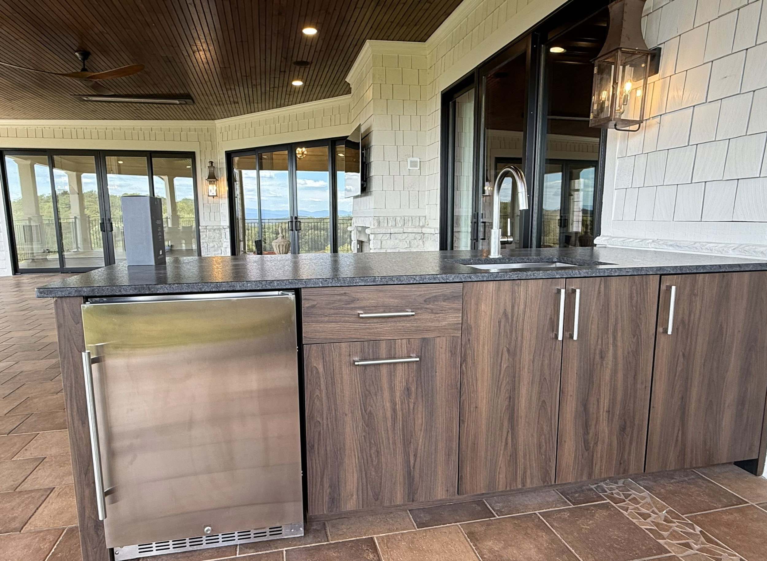 Outdoor kitchen counter with a wood cabinet, a stainless steel mini-fridge, and a sink with a modern faucet, with sliding glass doors and a view of mountains in the background.