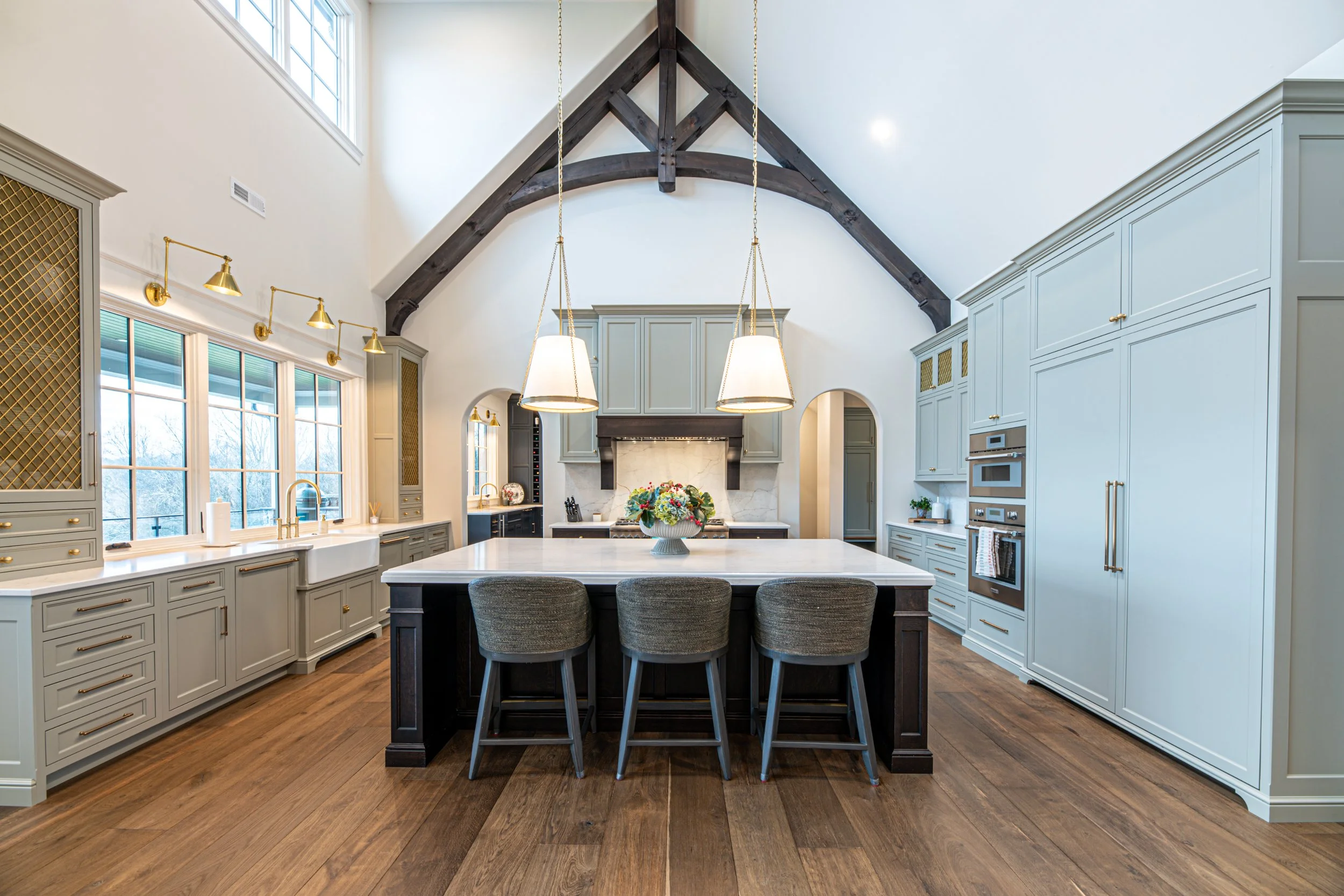 Modern kitchen with high vaulted ceiling, exposed dark wooden beams, light gray cabinetry, large center island with three upholstered chairs, hardwood flooring, and multiple lighting fixtures, including pendant lights and track lighting.
